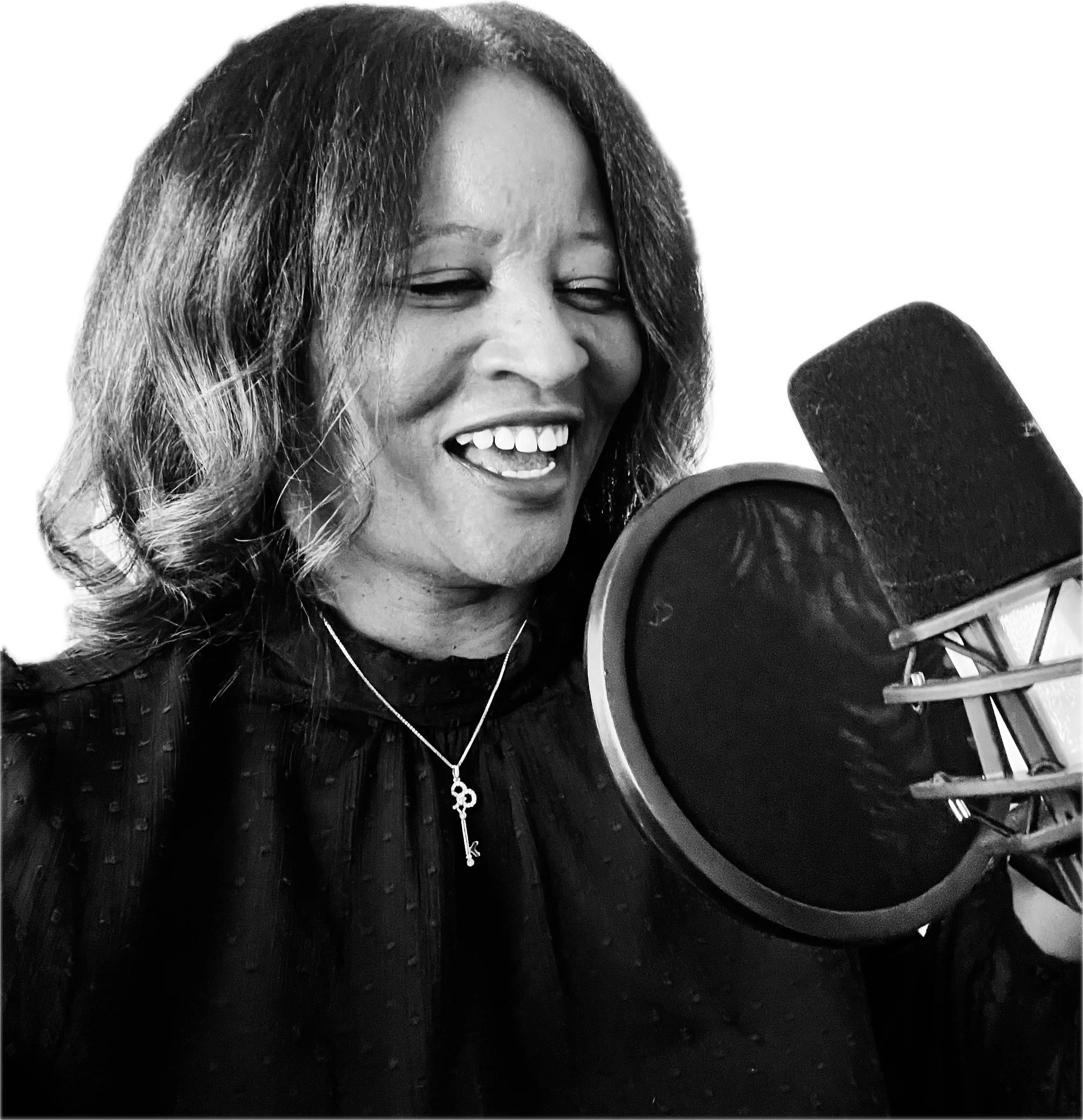 A woman with a great voice singing or speaking into a professional microphone in her home studio smiling with eyes closed, wearing a necklace with a key pendant, and a dark blouse with a textured pattern.