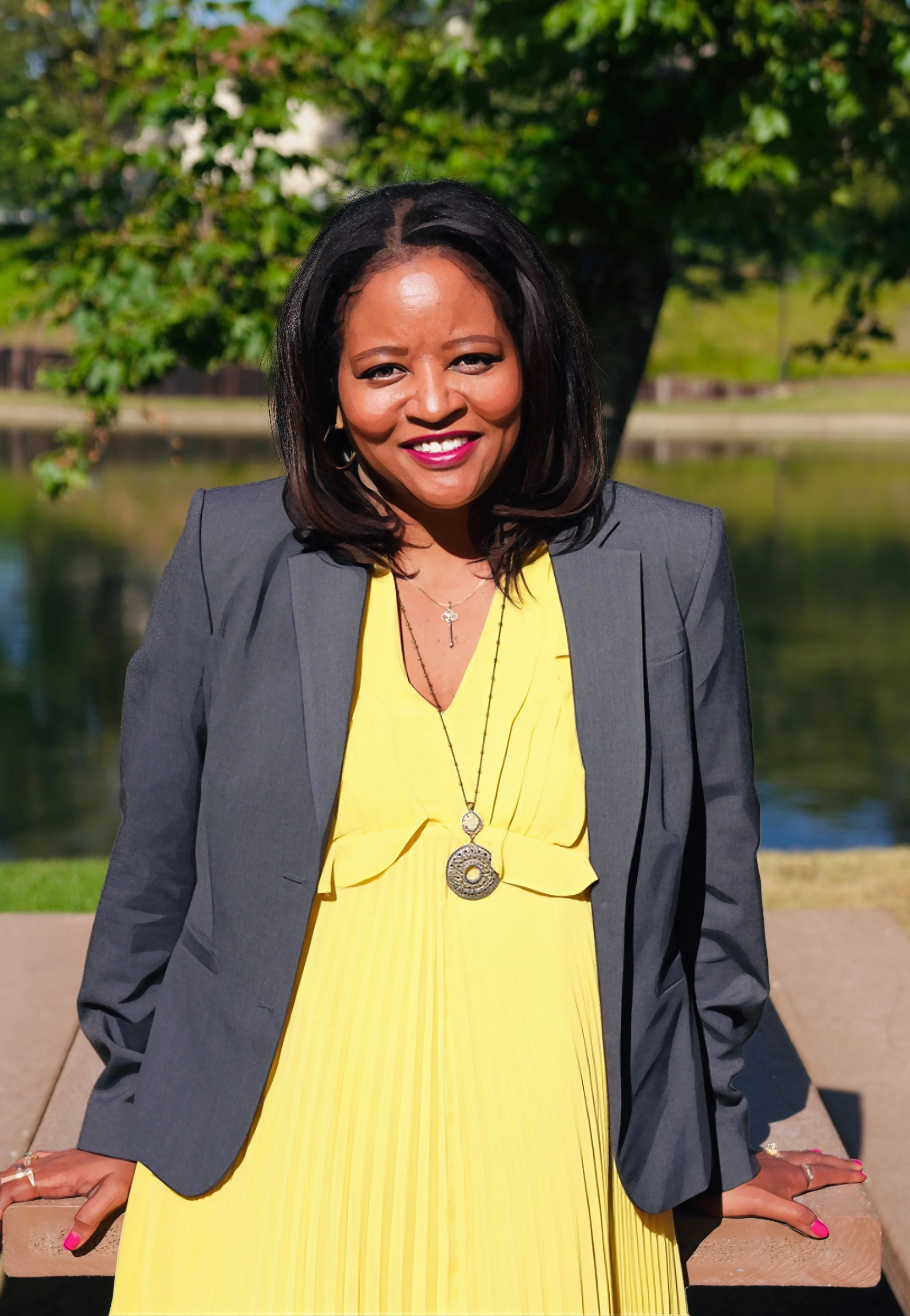 A woman with shoulder-length black hair and medium skin tone, smiling outdoors near a body of water, wearing a yellow dress, gray blazer, and jewelry. Calm. Serene. Relaxing.