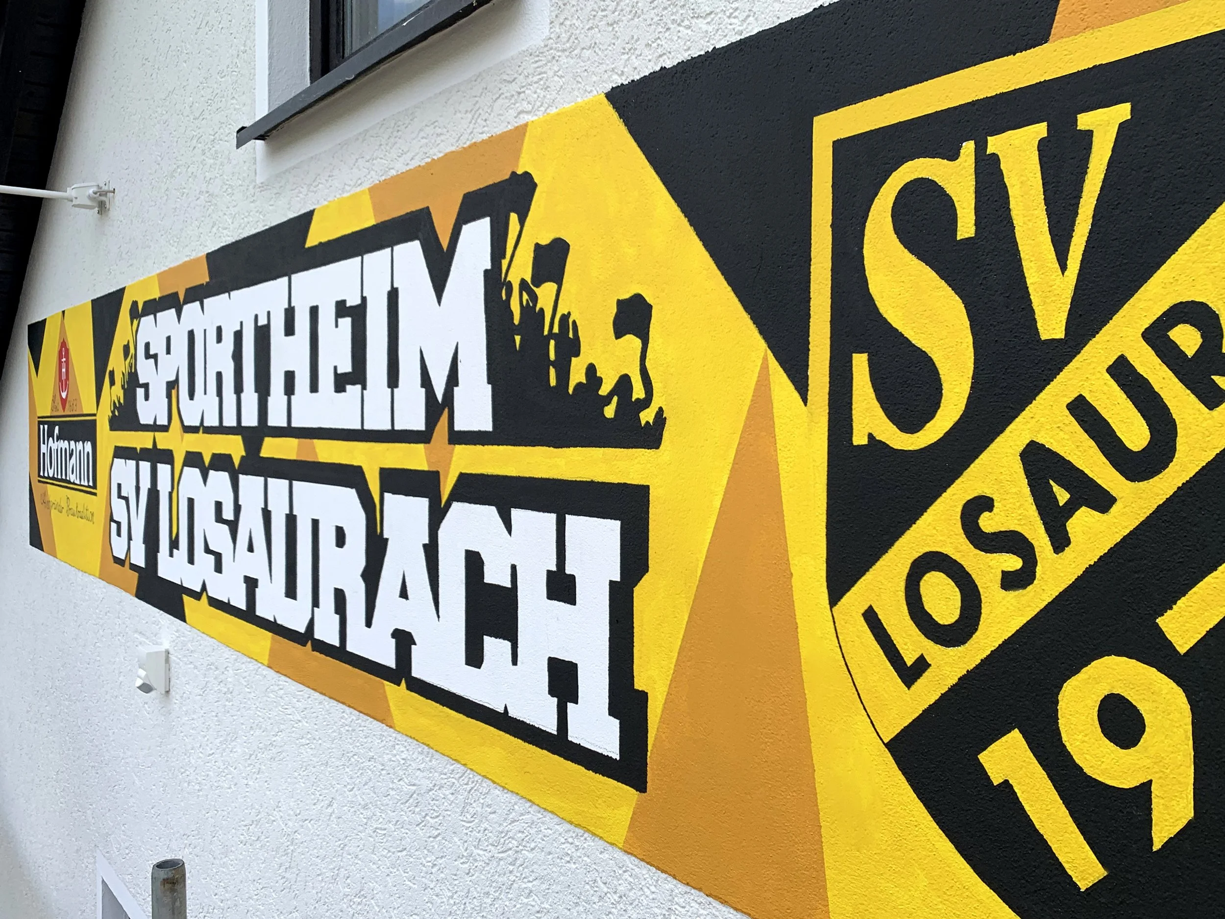 Großes Graffiti auf der Fassade eines weißen Hauses. Das Graffiti zeigt das Logo und den Namen des Sportvereins SV Losaurach, inklusive gelben Farben und weiteren Wappen in dynamischen Winkel.