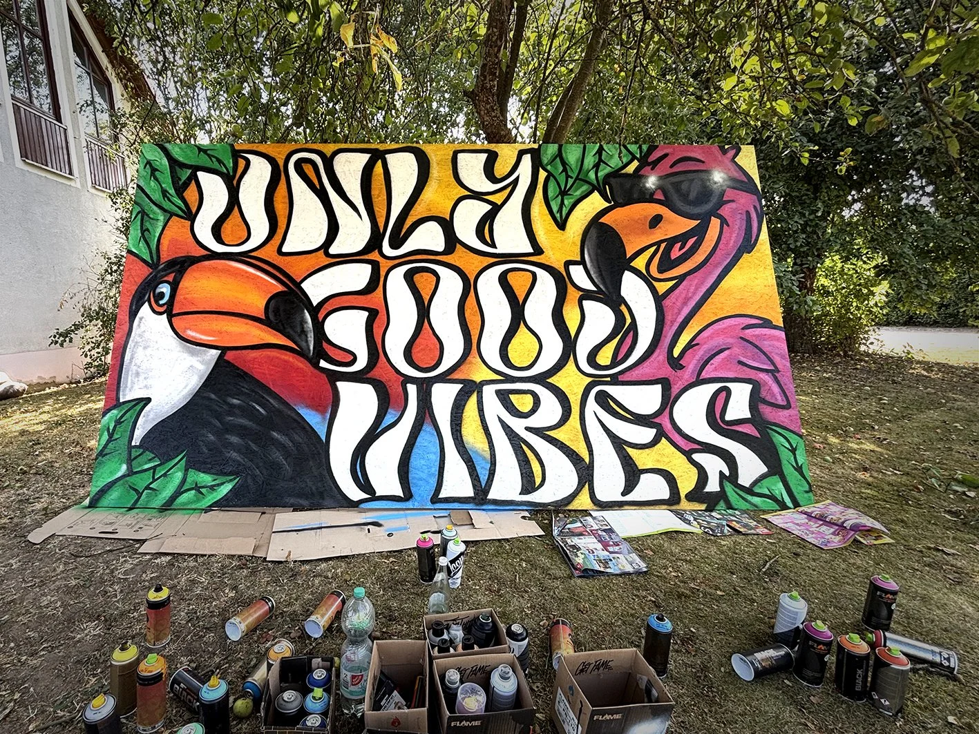 Ein buntes Graffiti mit Vögeln und umgebenden grünen Blättern, das auf einer großen Wand gemalt wird, mit Spraydosen und Malutensilien im Vordergrund. Zudem ist ein weißer Text zu sehen.