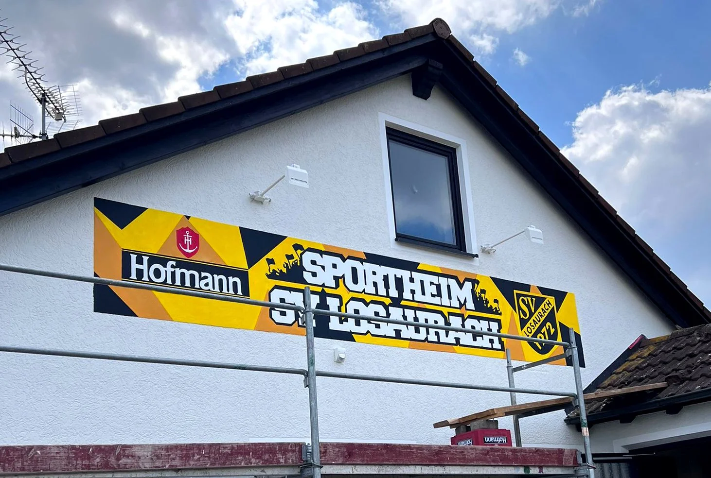 großes Graffiti auf der Fassade eines weißen Hauses. Das Graffiti zeigt das Logo und den Namen des Sportvereins SV Losaurach, inklusive gelben Farben und weiteren Wappen.