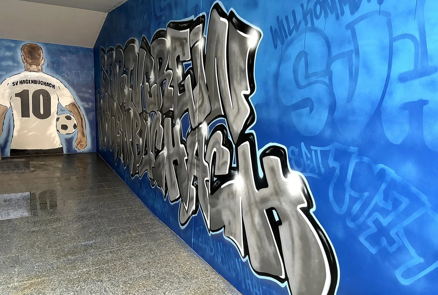 Graffiti-Kunstwerk mit grauer Schrift an einer Wand mit blauer Hintergrundfarbe, das einen Fußballspieler mit Trikot Nummer 10 und dem Schriftzug Hagenbüchach zeigt, sowie weiteren Graffiti-Texten.