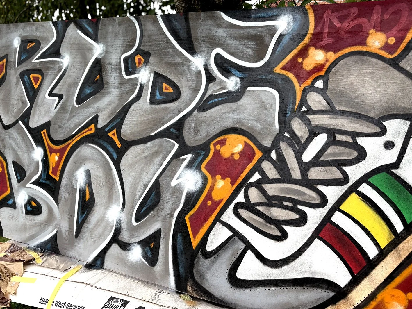 Graffiti-Kunstwerk mit grauen Buchstaben, einem Turnschuh und bunten Elementen auf einer Wand.