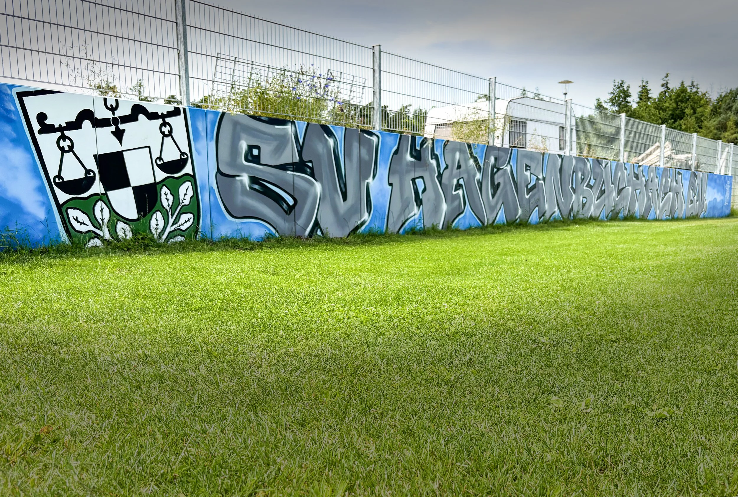 Graffiti an einer blauen Mauer am Spielfeldrand, mit einem Wappen des Vereins und großen grauen Buchstaben.