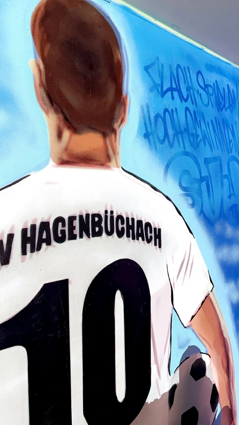 Rückansicht eines Spielers beim Fußball, Trikot mit Nummer 10 und Namen 'HAGENBÜCHACH', im Hintergrund eine blaue Wand mit Graffiti