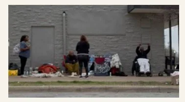 Four women standing and sitting on a sidewalk outside a brick building, some with items on the ground and a small table with a cloth.