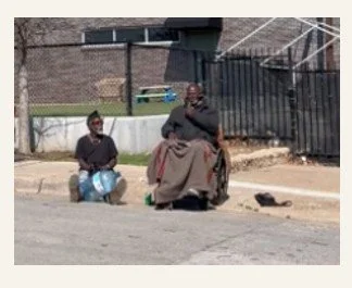 Two men sitting on the sidewalk; one is playing a drum, and the other is sitting with arms crossed, near a fence and a building.