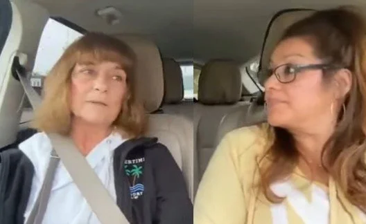 Two women sitting in the backseat of a car, engaging in a conversation. One woman has shoulder-length blonde hair and is wearing a white shirt with a black jacket, while the other woman has long brown hair, glasses, and a yellow top.