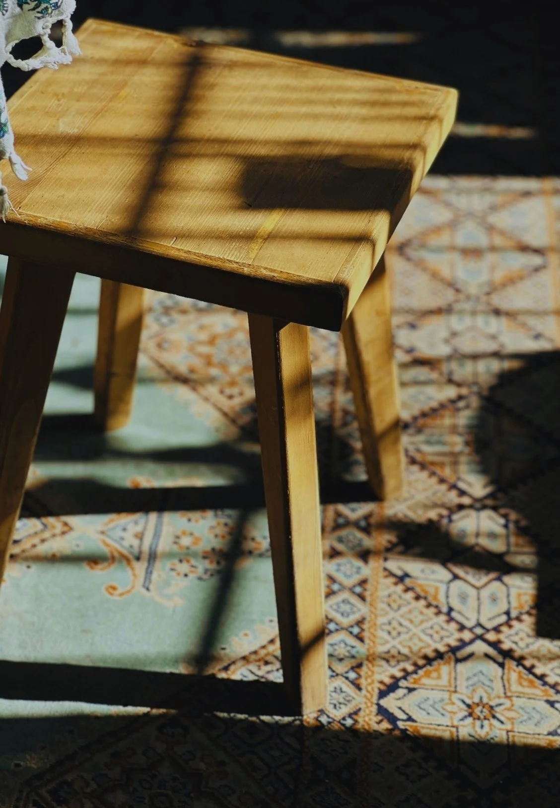 Close-up of a wooden stool casting shadows on a patterned rug with sunlight streaming through window shadows.