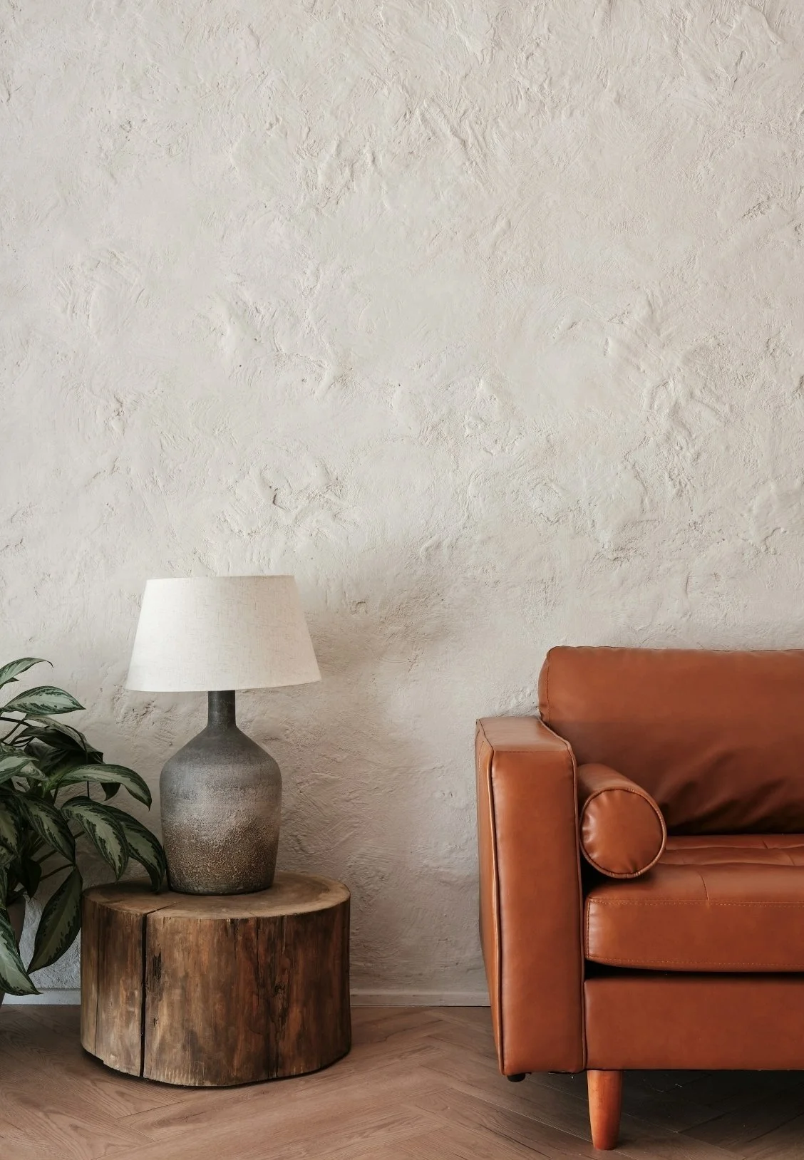 Living room corner with a brown leather sofa, a wooden side table with a large ceramic lamp, and a green potted plant against a textured white wall.