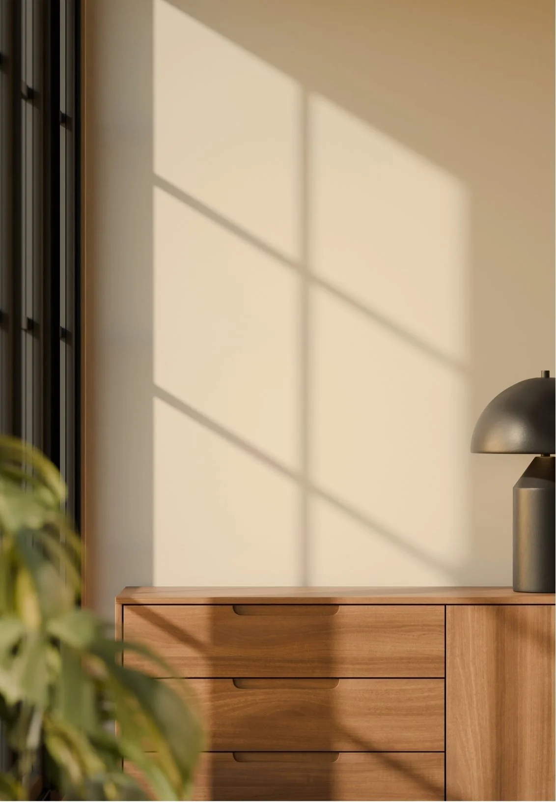 Sunlight casting windowpane shadows on a beige wall above a wooden dresser with a black lamp and green plant leaves in the foreground.