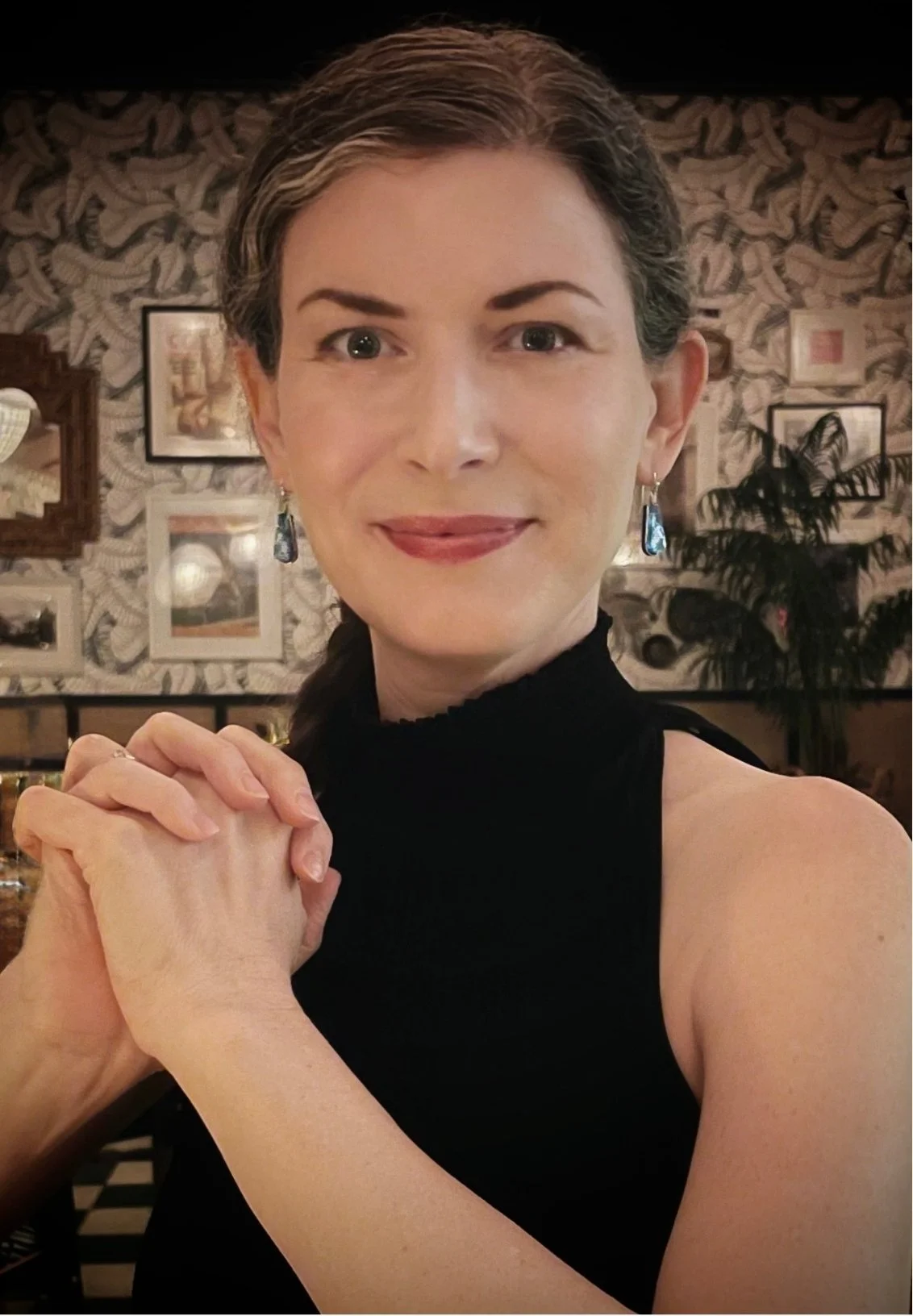 Lauren Maher, therapist in Los Feliz, CA,  wearing a black top, sitting in a room with patterned wallpaper and framed pictures, smiling at the camera with her hands clasped.