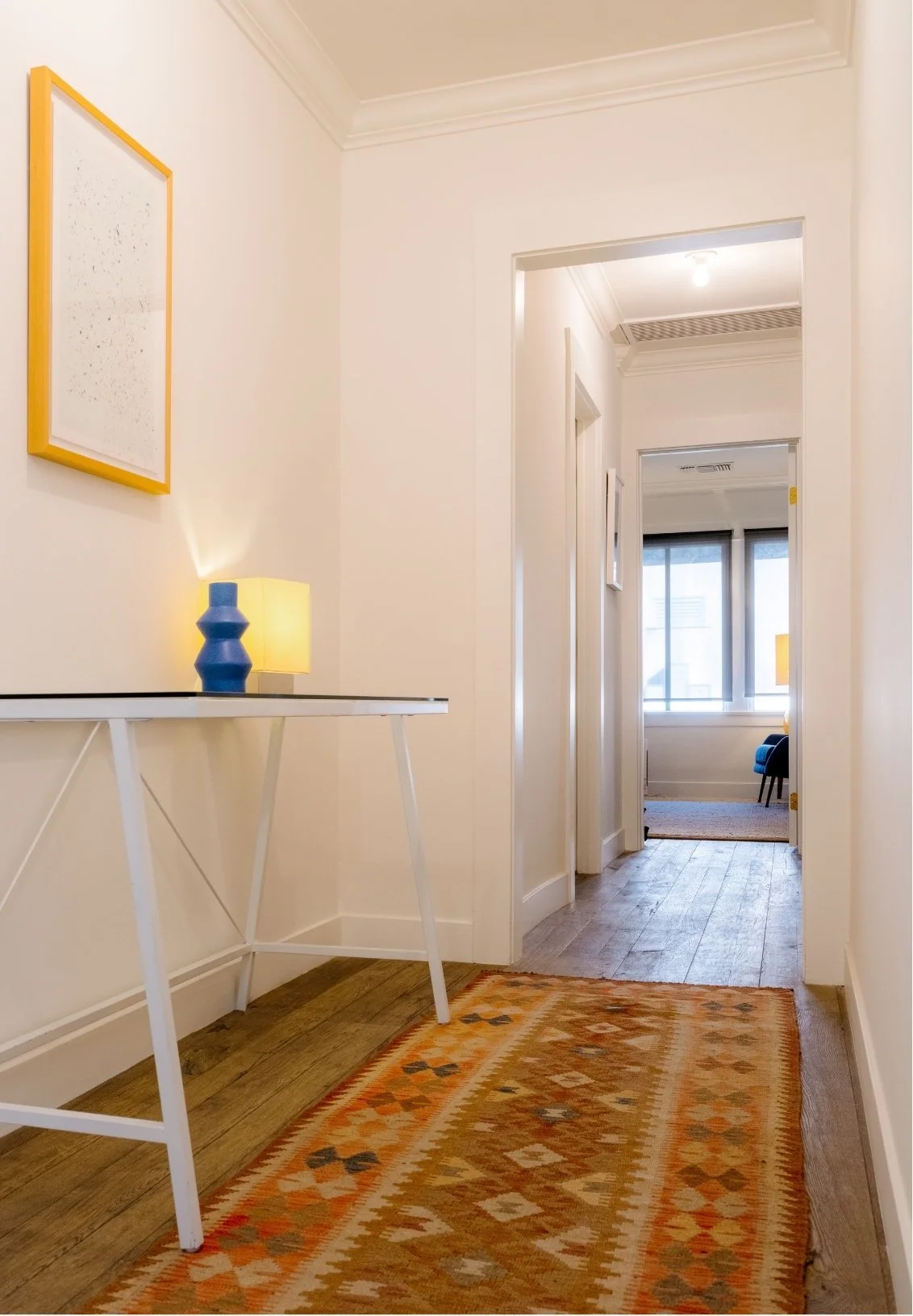 A hallway with a colorful patterned rug on hardwood floor, a white table with a blue vase and yellow lamp, framed artwork on the wall, view into a room with large window and blue armchair.