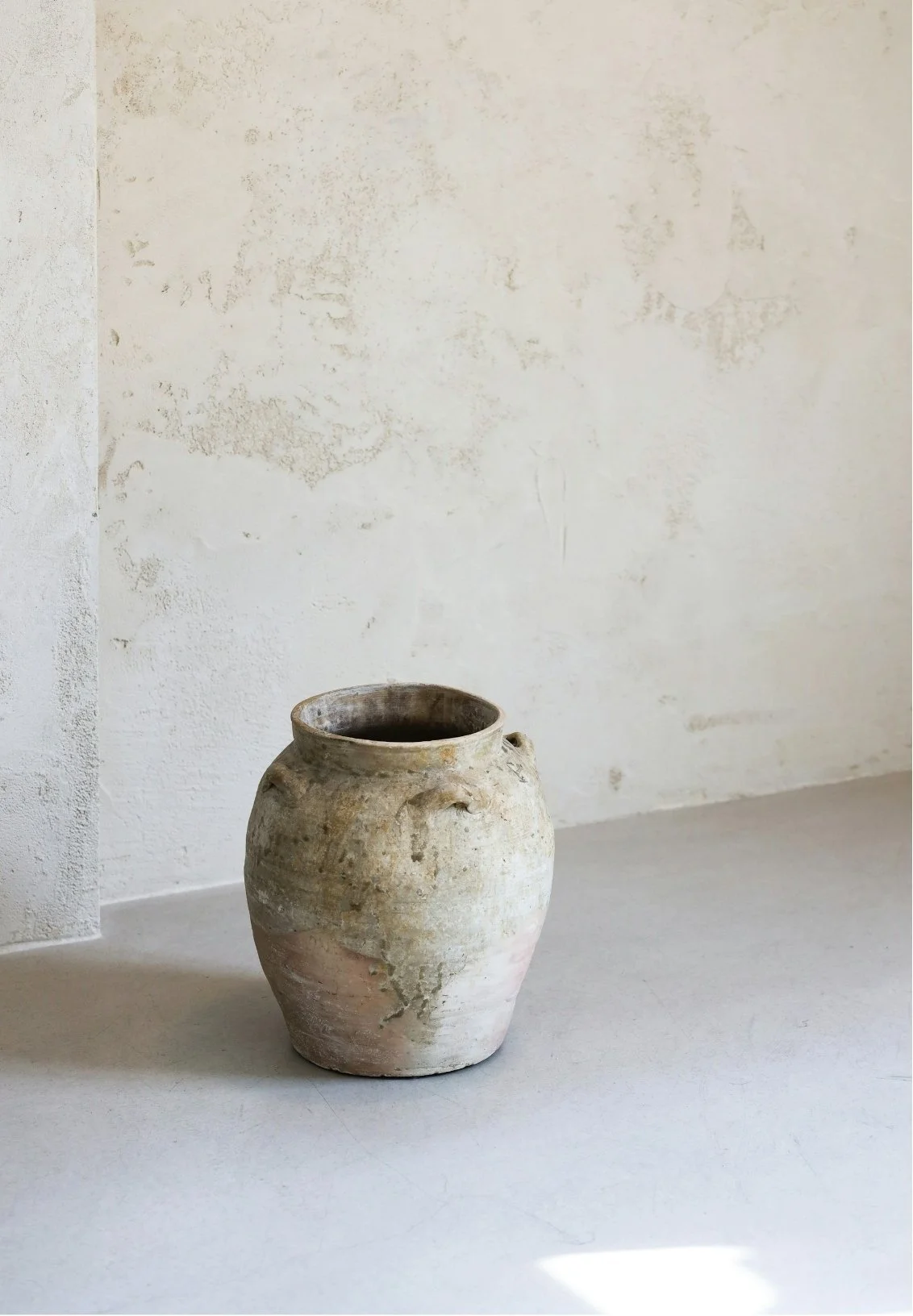 An old, weathered clay pot sitting on a white surface against a textured, off-white wall.
