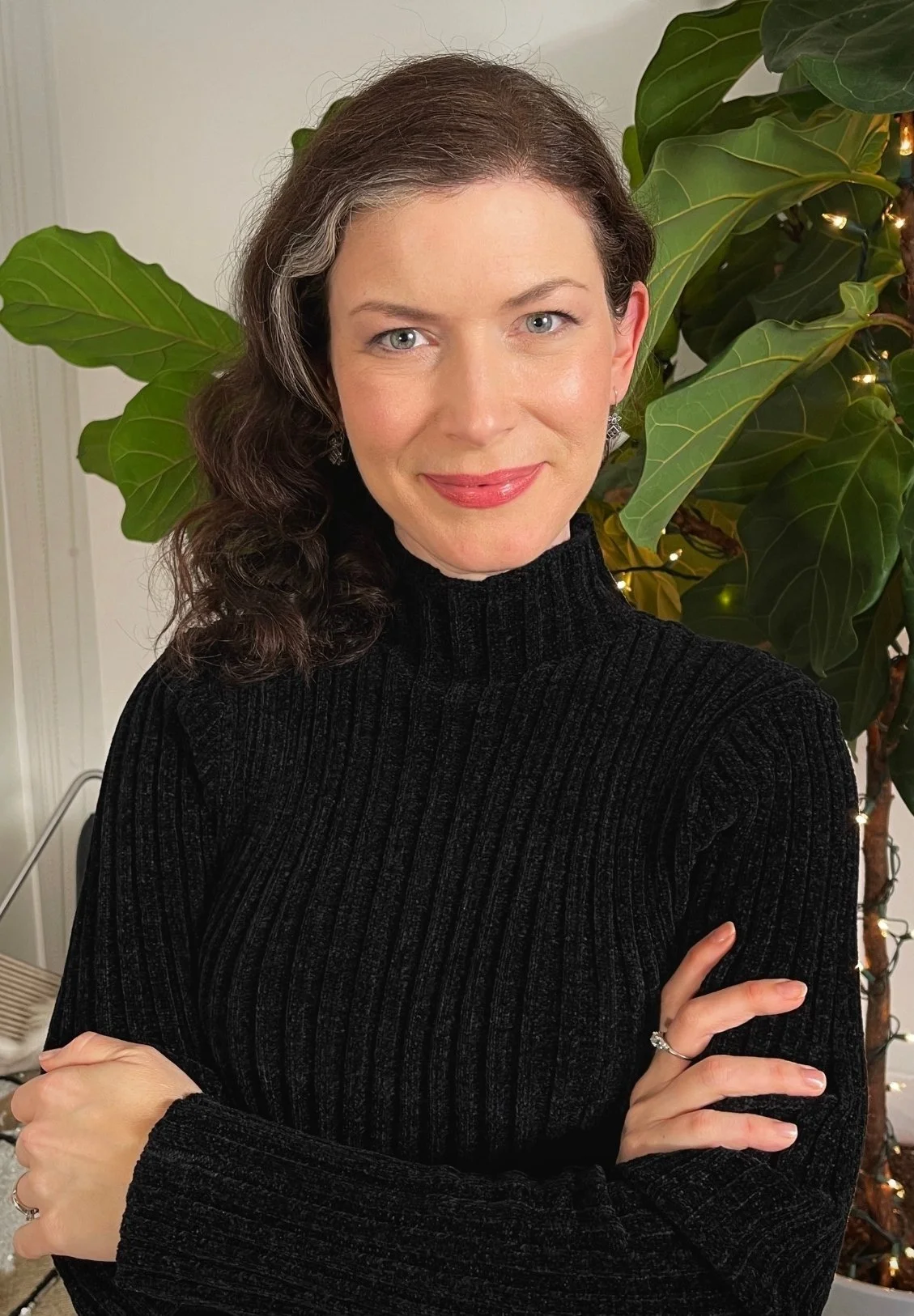 Lauren Maher, therapist in Los Feliz, CA, smiling, wearing a black ribbed turtleneck sweater, standing in front of large green leaves with string lights.