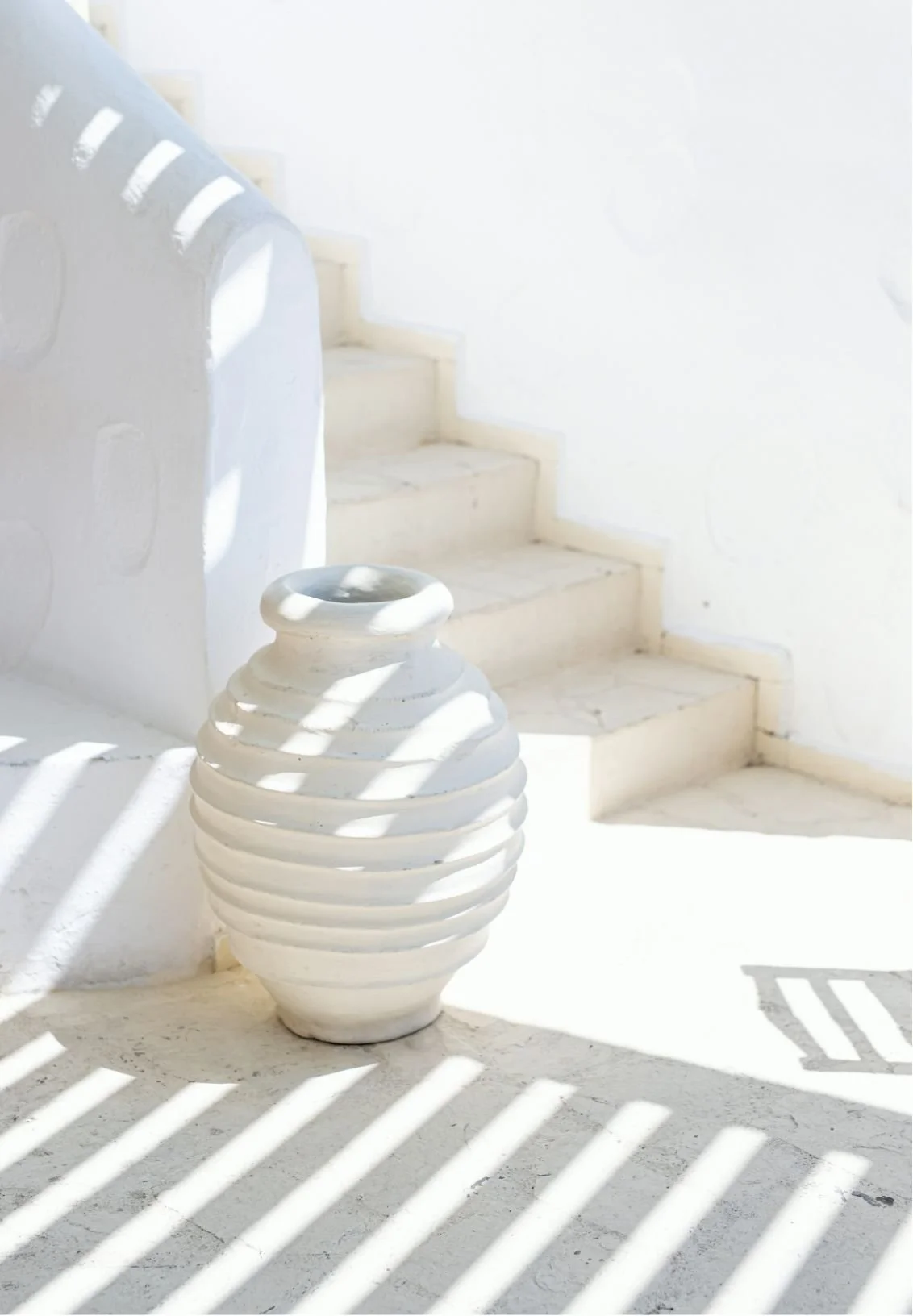 White stairway with a large white ceramic vase in the foreground, casting striped shadows on the floor, with a white wall in the background.