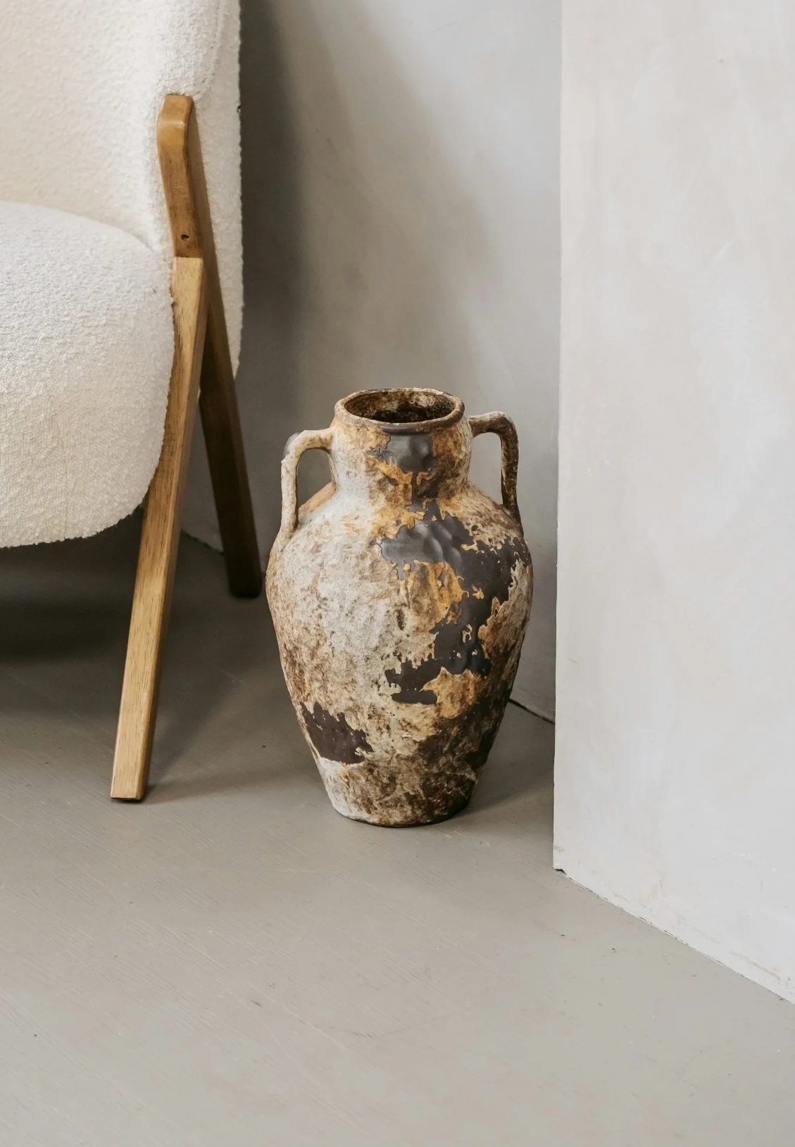 An ancient, weathered ceramic vase with two handles, placed on a light-colored floor near a corner of a wall. Part of a chair with wooden legs is visible on the left.