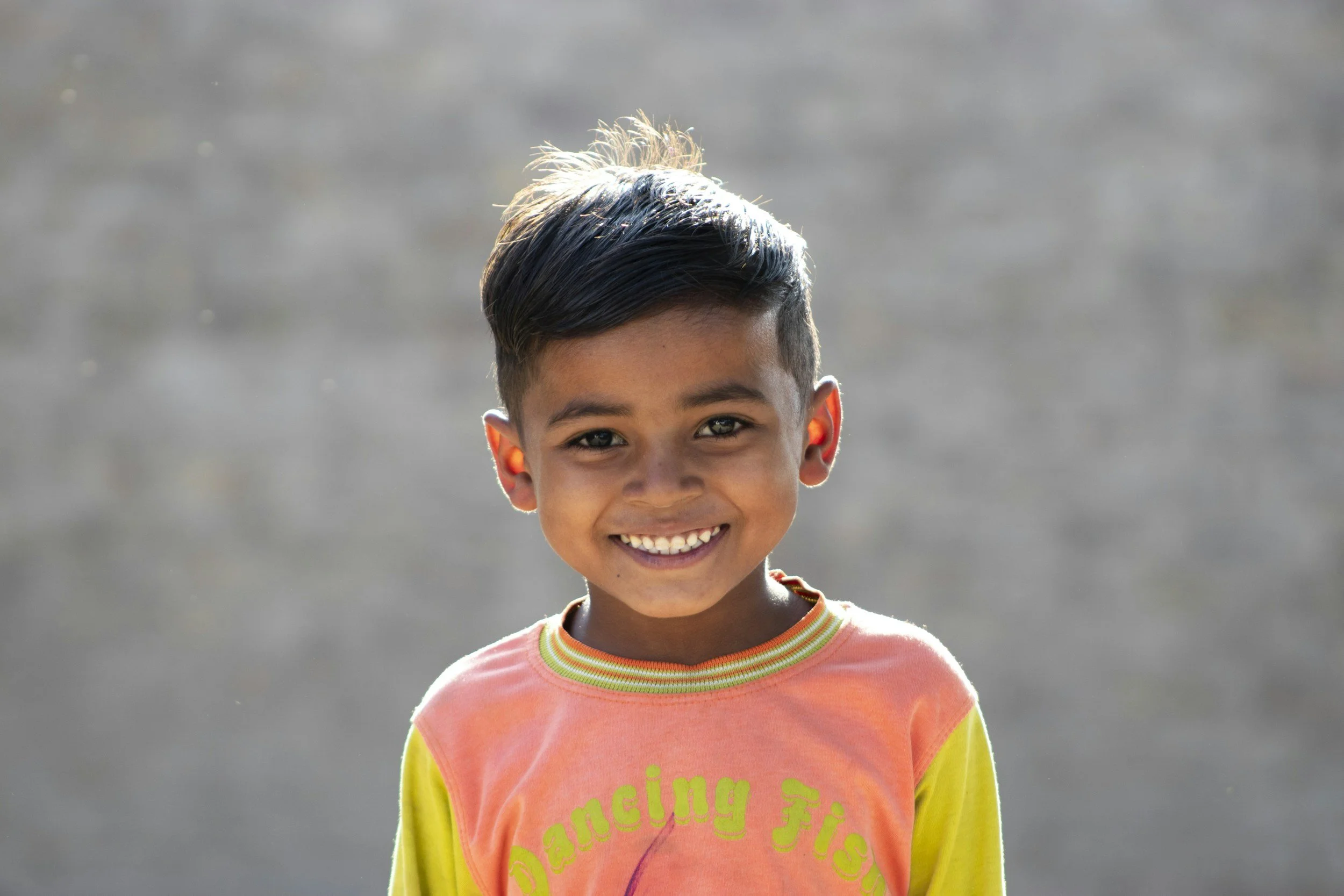 A young boy with dark hair and brown skin smiling at the camera, wearing a colorful shirt with the words 'Dancing Fish' on it, standing against a blurred outdoor background.