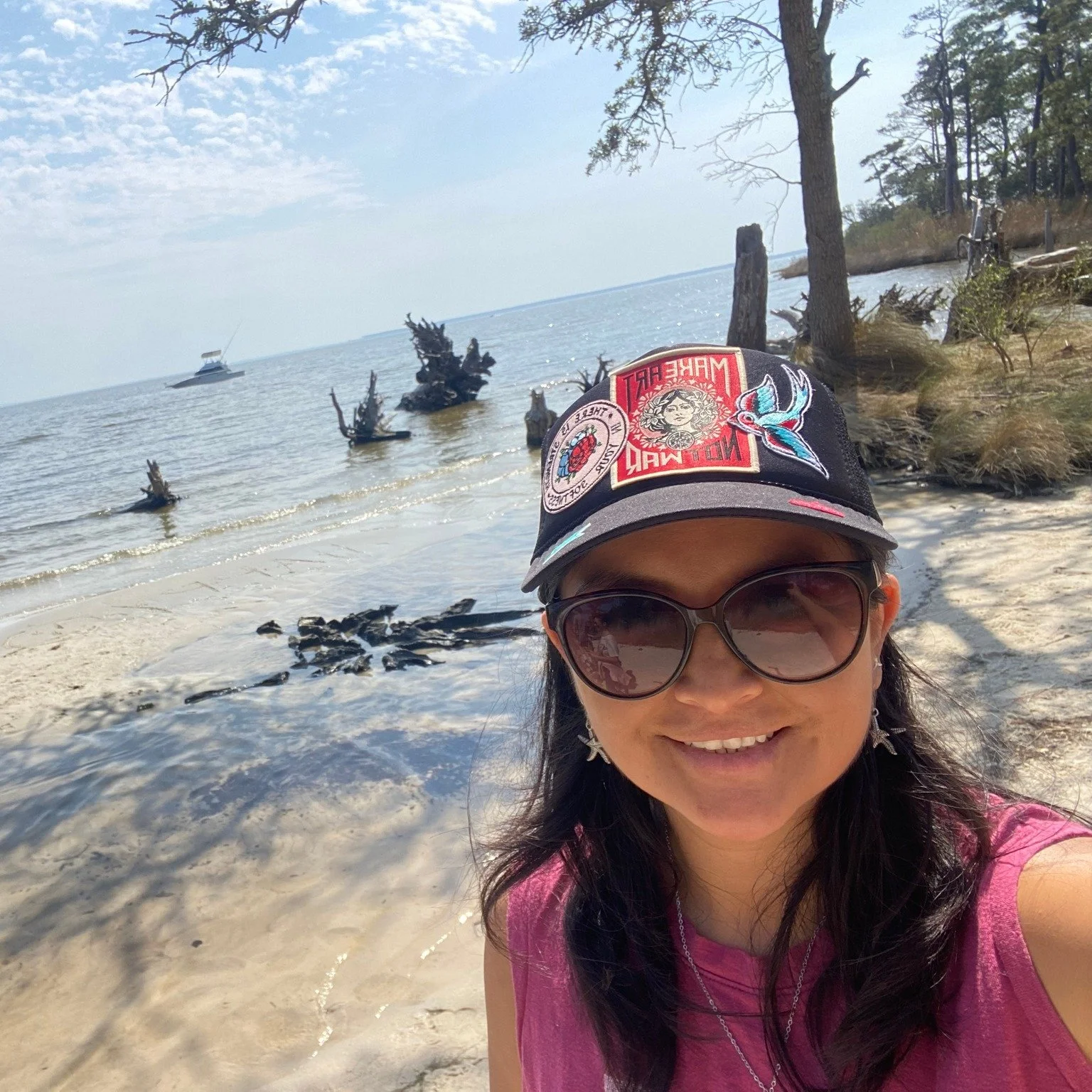 A woman wearing sunglasses and a black cap with colorful patches takes a selfie on a sandy beach with driftwood and trees, with a boat on the water in the background.