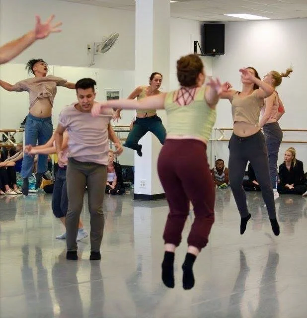 Group of dancers practicing in a dance studio, some jumping and others standing, with a mirror and audience seated in the background.