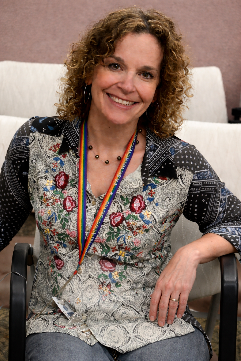 A woman with curly hair and a colorful or patterned blouse, smiling while sitting on a chair. She has a rainbow-colored lanyard around her neck.