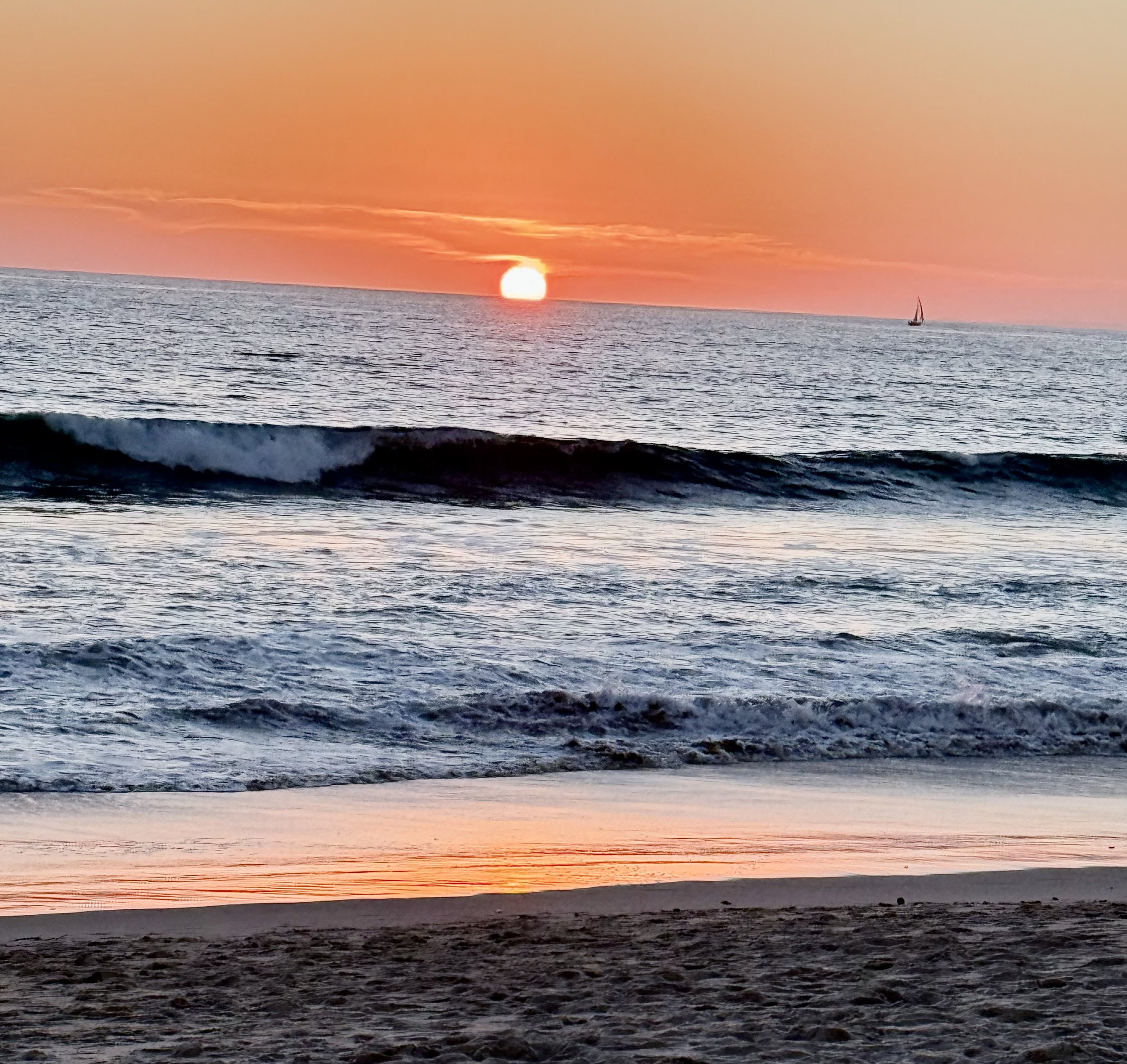 Sunset over the ocean with waves crashing on the shore and a sailboat in the distance.