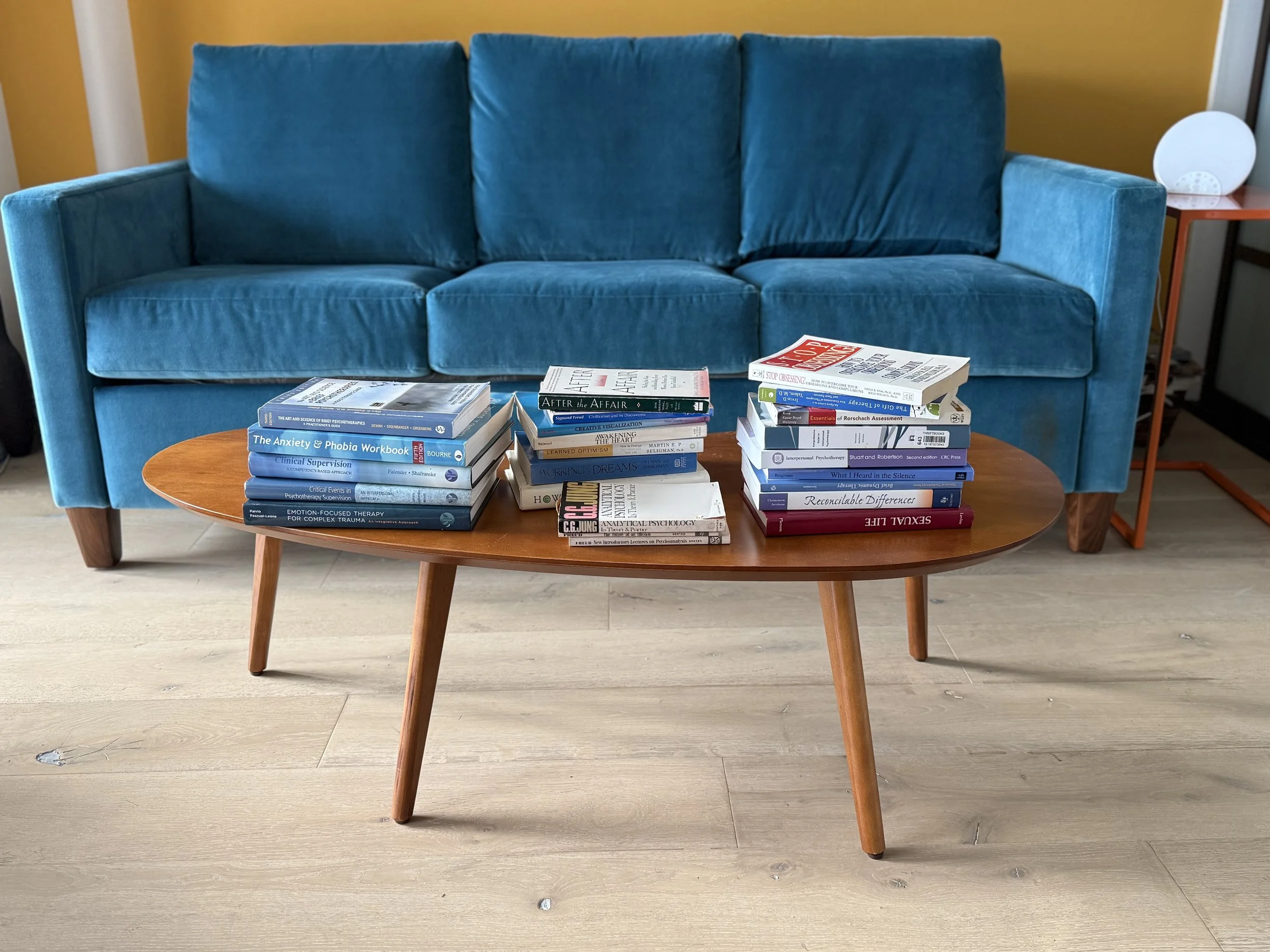 A blue sofa with a wooden coffee table in front of it, holding stacks of books related to psychology, therapy, and mental health. There is a side table with a round white lamp beside the sofa.