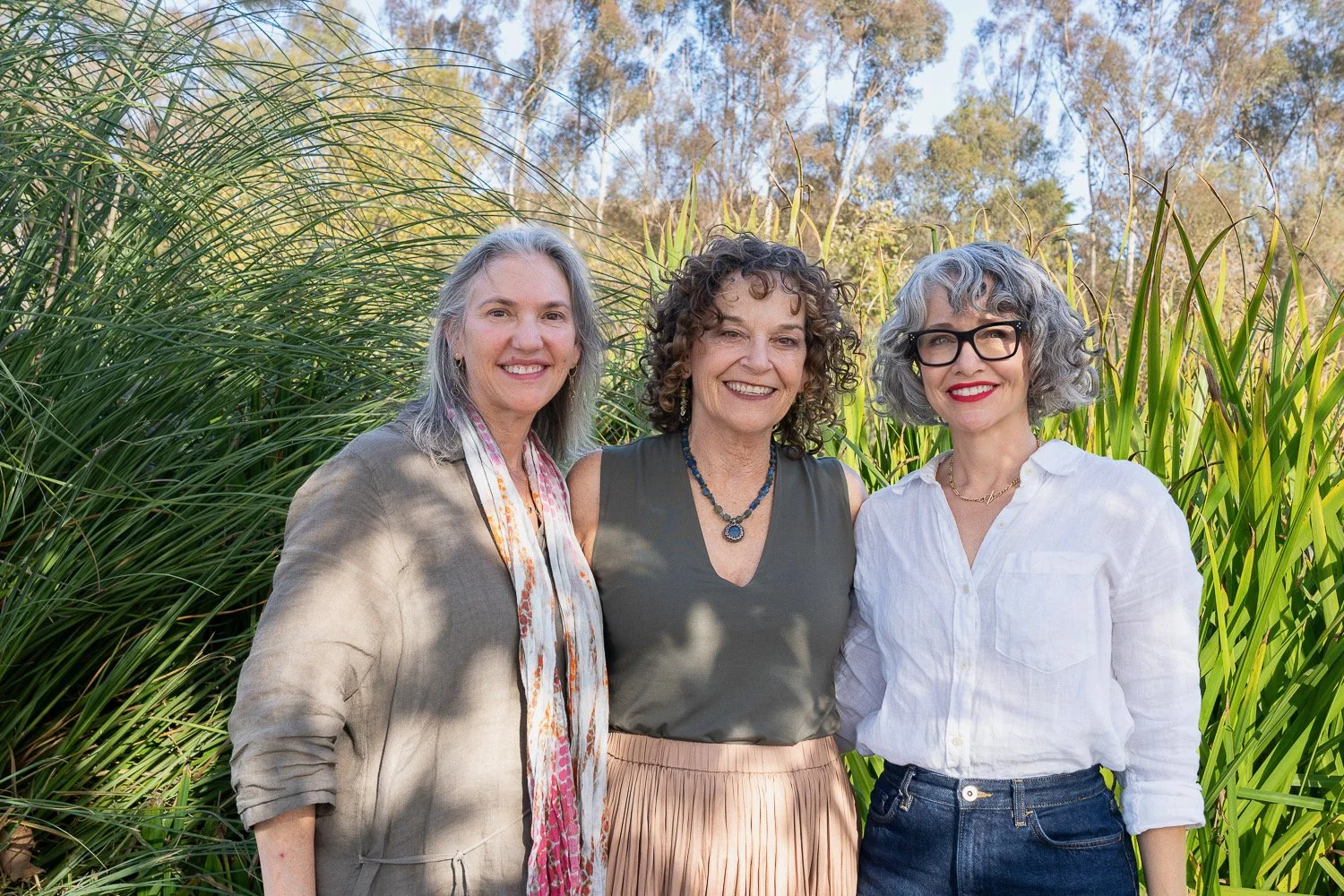 Three women standing outdoors in front of tall grass and trees, smiling at the camera.