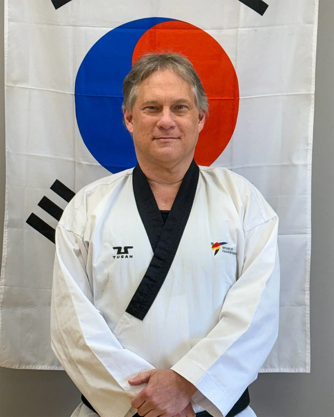 John Salchow, a caucasian man with grey hair, is wearing a Taekwondo uniform and standing in front of a Korean flag.