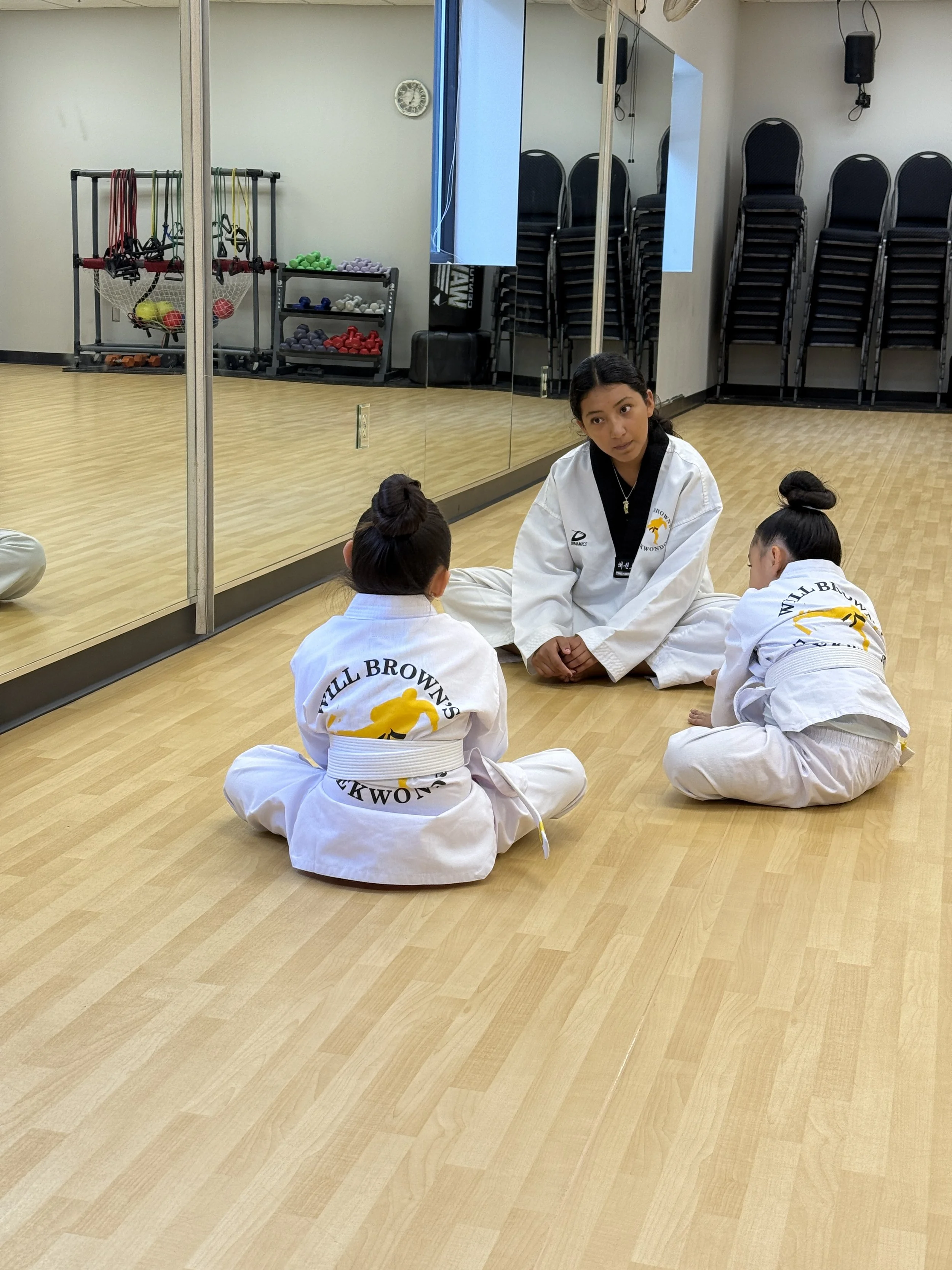 In Will Brown's Taekwondo school, assistant instructor Danielle is sitting on the floor, talking to two young girls in white uniforms, in front of a mirrored wall with stacked chairs, fitness equipment, and a large mirror.