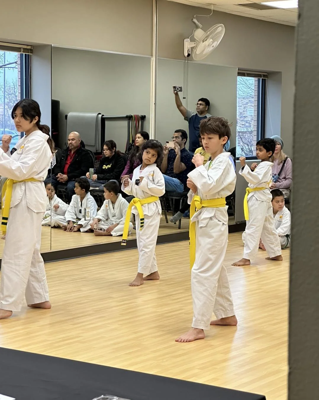 Young students in gold belts performing during a belt testing at Will Brown's Taekwondo, with family members watching in the background.