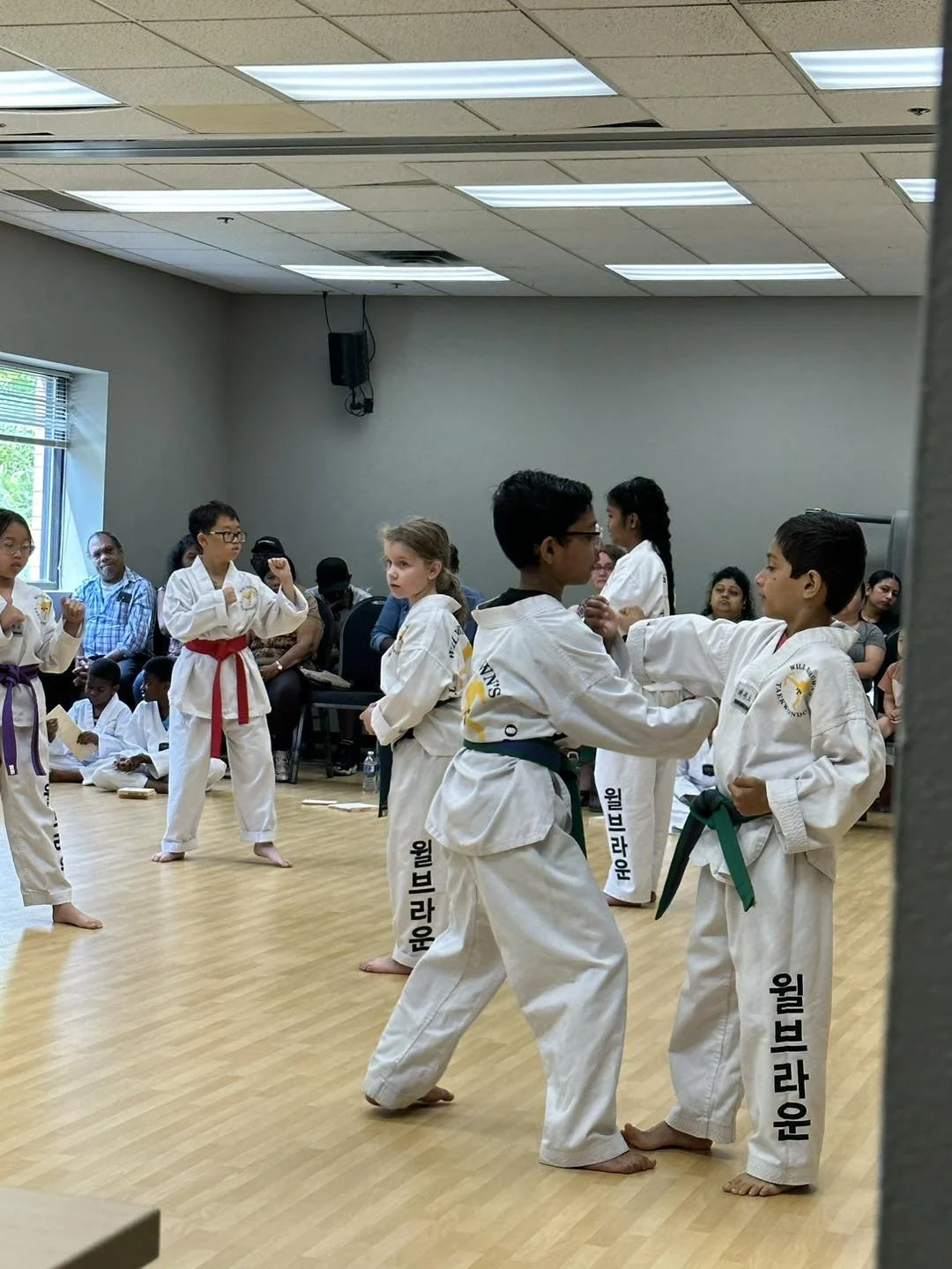 Students practicing one-steps during a belt testing at Will Brown's Taekwondo, with family members watching in the background.