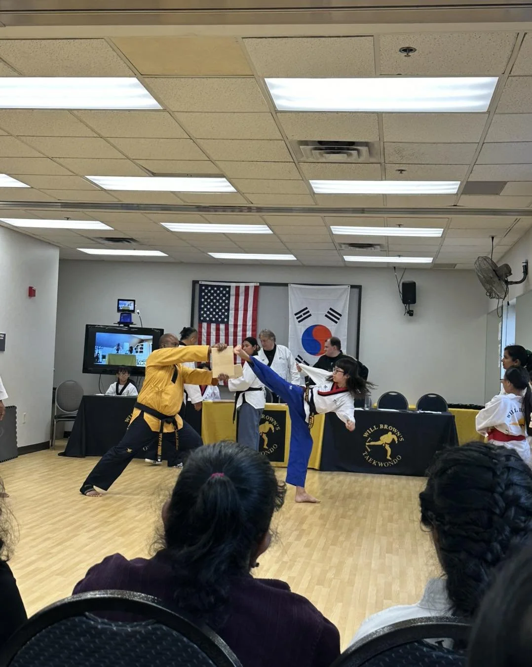 A student executes a high kick toward a board held by Master Will Brown during a belt testing at Will Brown's Taekwondo, with family members watching in the background.