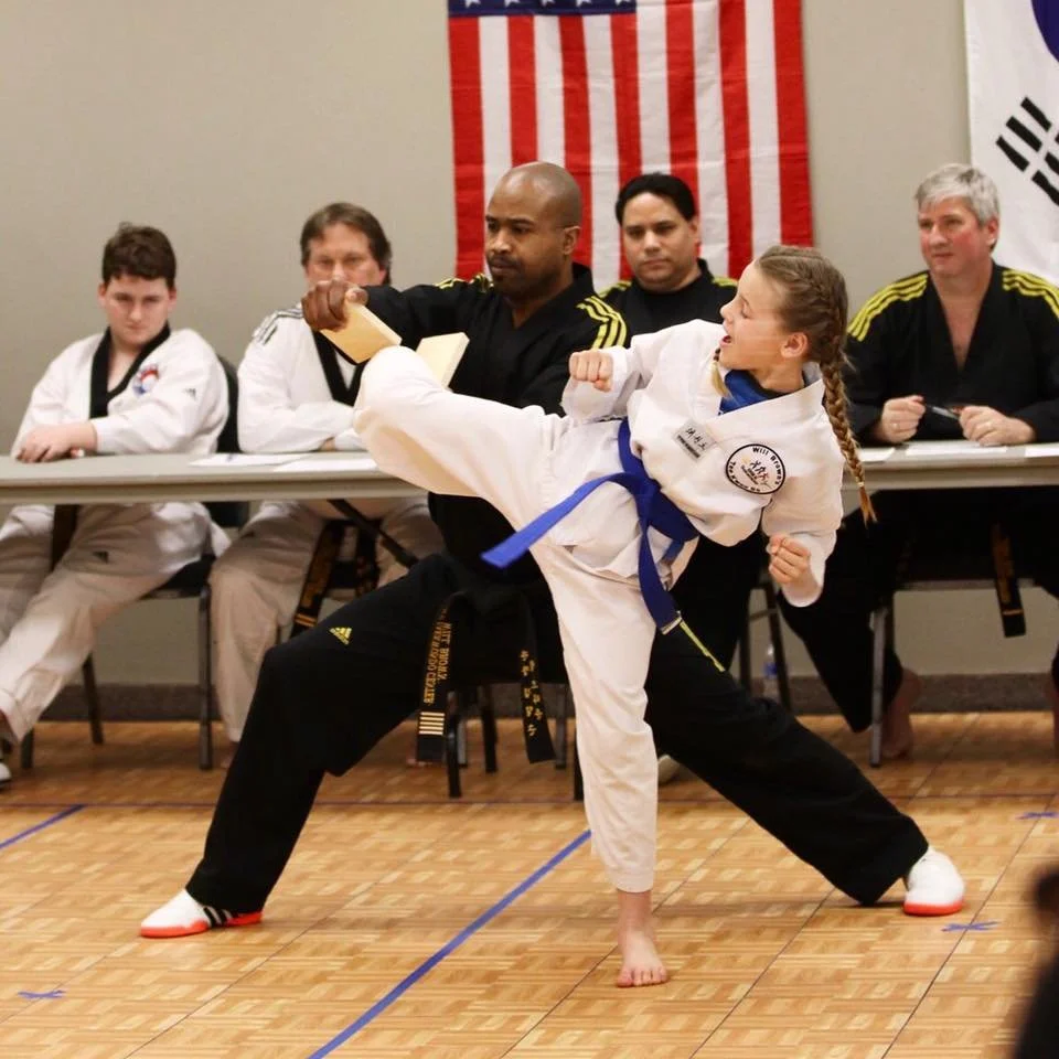 A student executing a high kick toward a board held by Master Will Brown during a belt testing at Will Brown's Taekwondo, with family members watching in the background.
