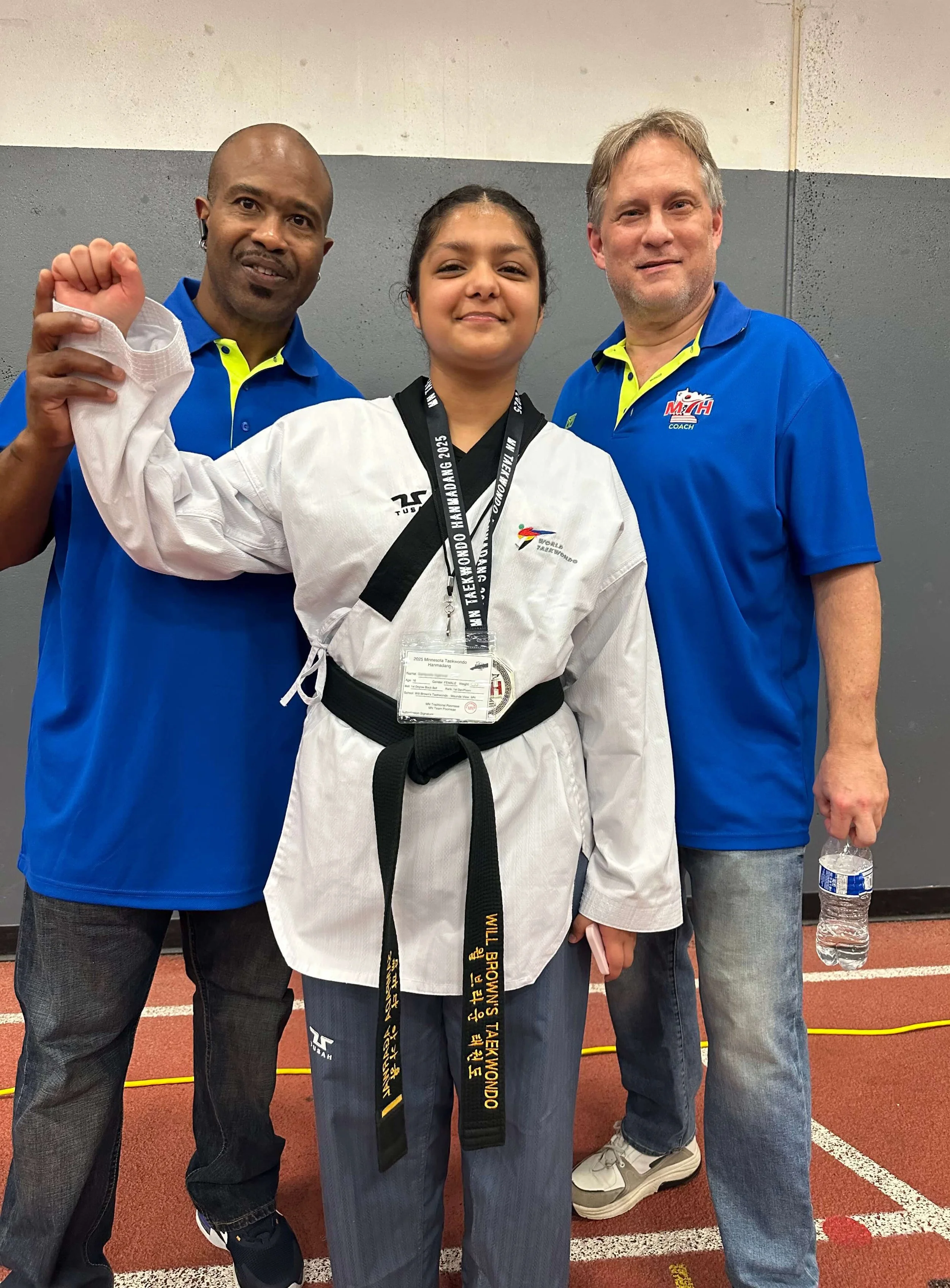 Master Will Brown and Lead Instructor John Salchow celebrating with a young competitor in a white taekwondo uniform and black belt at a tournament.