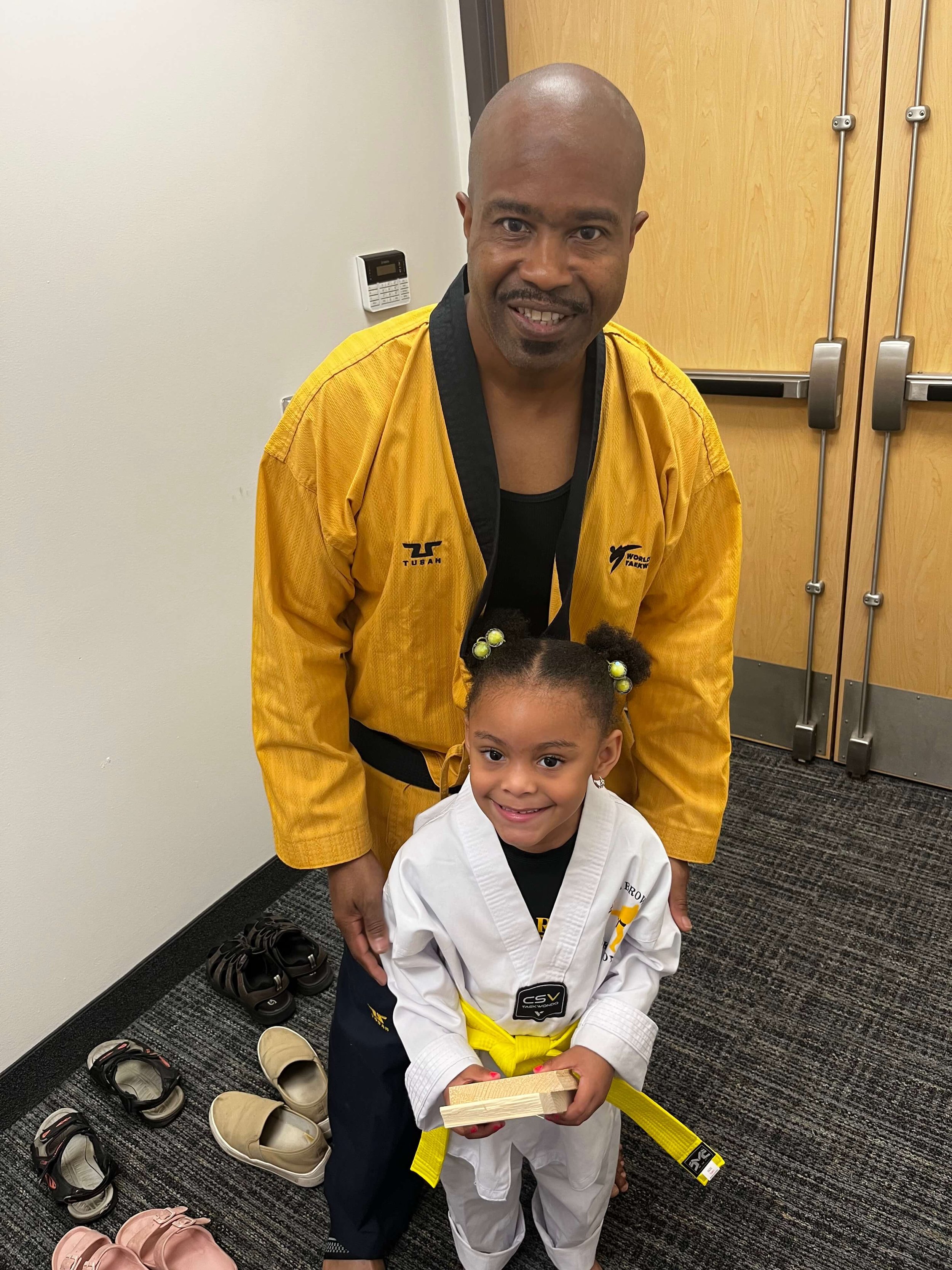 Master Will Brown poses with a young student who just earned her gold belt. They are both smiling, and the student is holding the pieces of the board she broke during her belt testing.