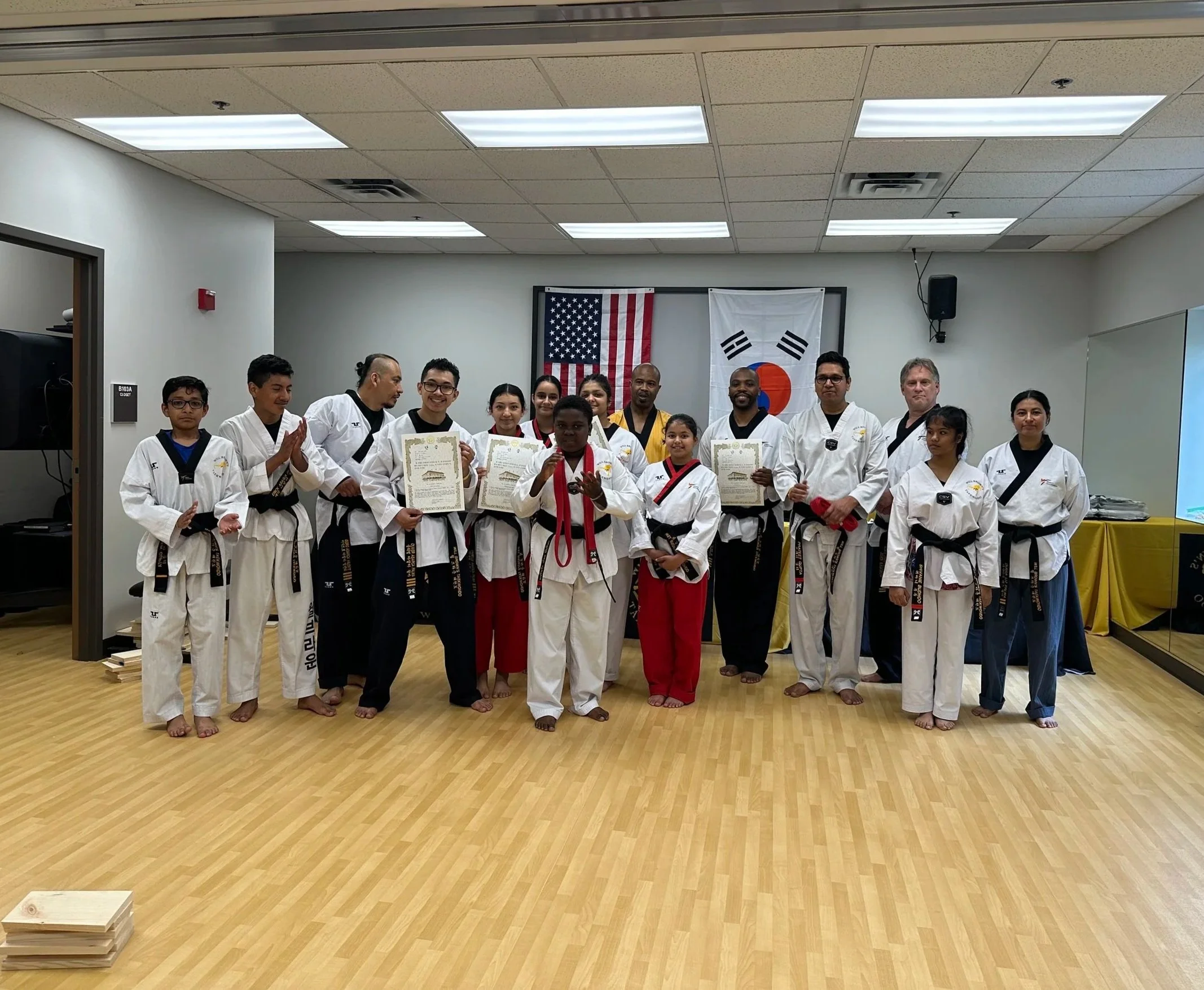 Master Will Brown and a group of students and instructors at Will Brown's Taekwondo pose together after a belt testing ceremony. Several students are holding certificates, with the American and South Korean flags displayed in the background.