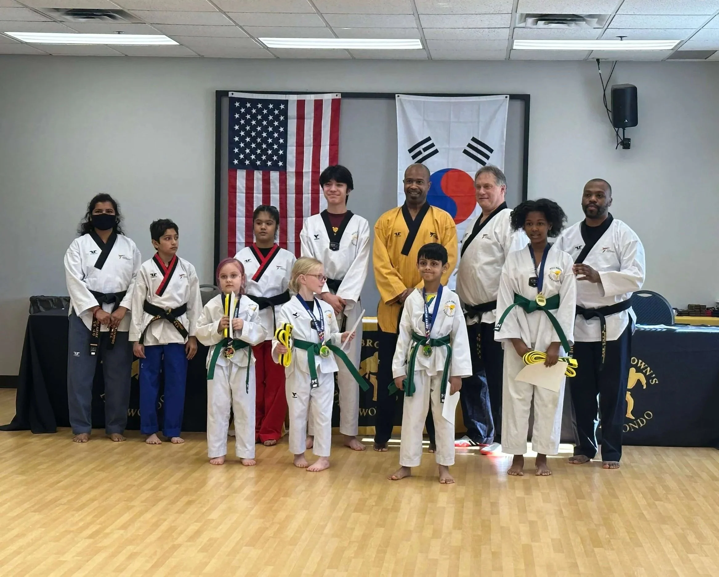 Master Will Brown and the instructors and students of Will Brown's Taekwondo pose together after a belt testing ceremony. Several students are wearing medals and holding trophies, with the American and South Korean flags displayed in the background.
