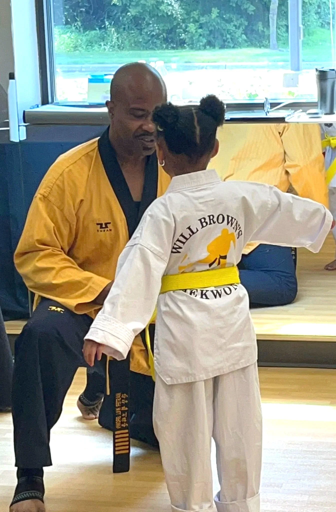 Master Will Brown is crouching down and helping tie a young student's newly earned gold belt.
