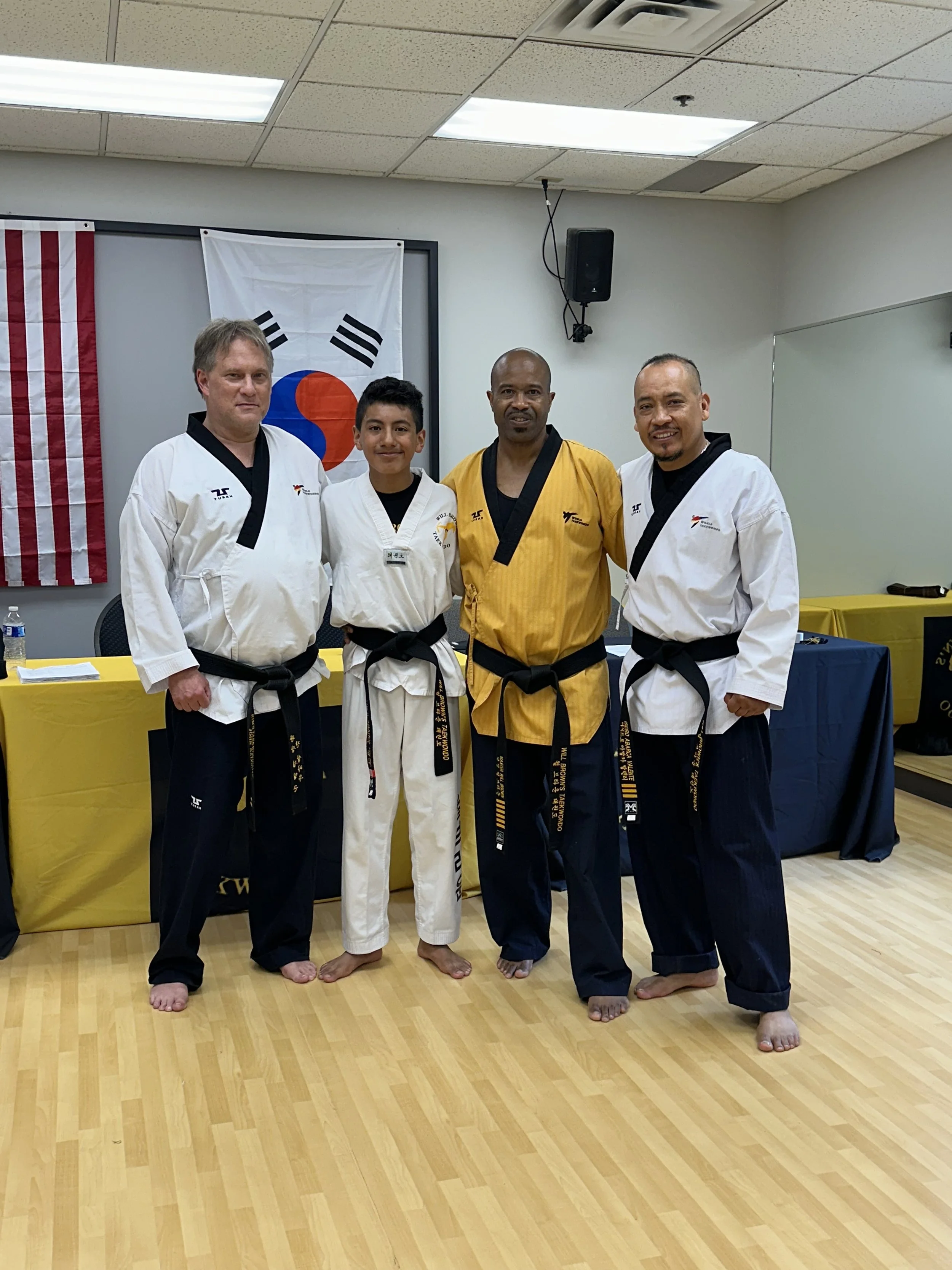 Master Will Brown and Lead Instructor John Salchow are standing with two students in Will Brown's Taekwondo, with South Korean and American flags in the background. They are wearing traditional uniforms and smiling.