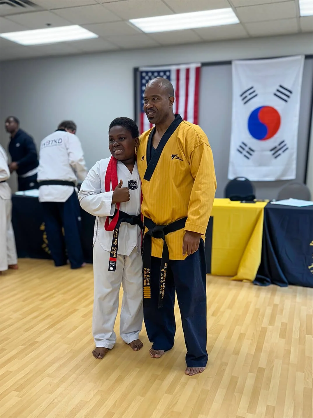 Master Will Brown posing with a student who just earned his black belt. The student is giving a thumbs up and they're both smiling. The American and South Korean flags are displayed in the background.