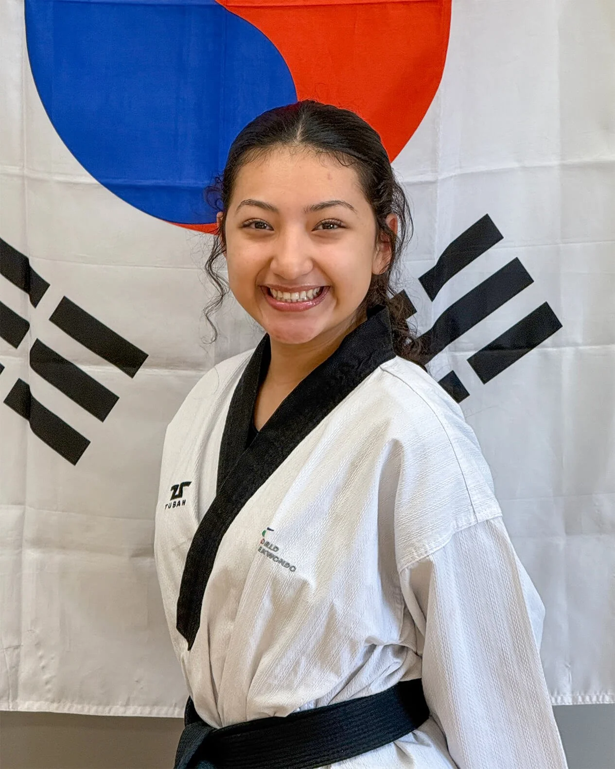 Danielle Abarca, a young woman with dark hair, is smiling, wearing a Taekwondo uniform, and standing in front of a Korean flag.