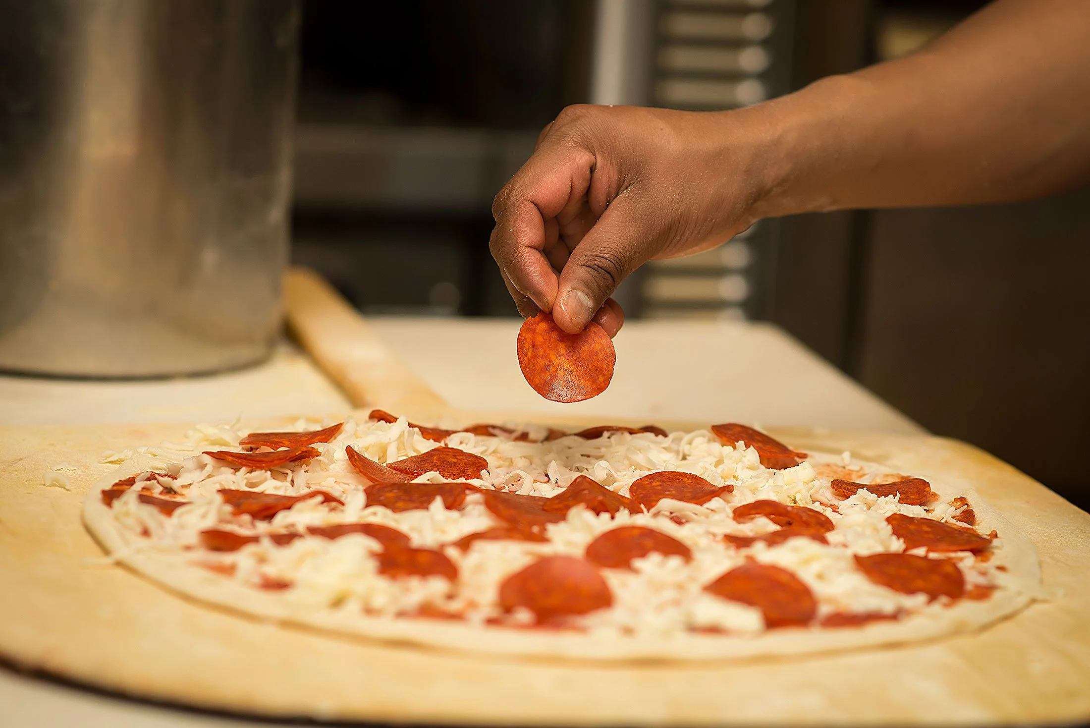 Close-up of a hand placing a slice of pepperoni on a pizza with cheese on a wooden surface.