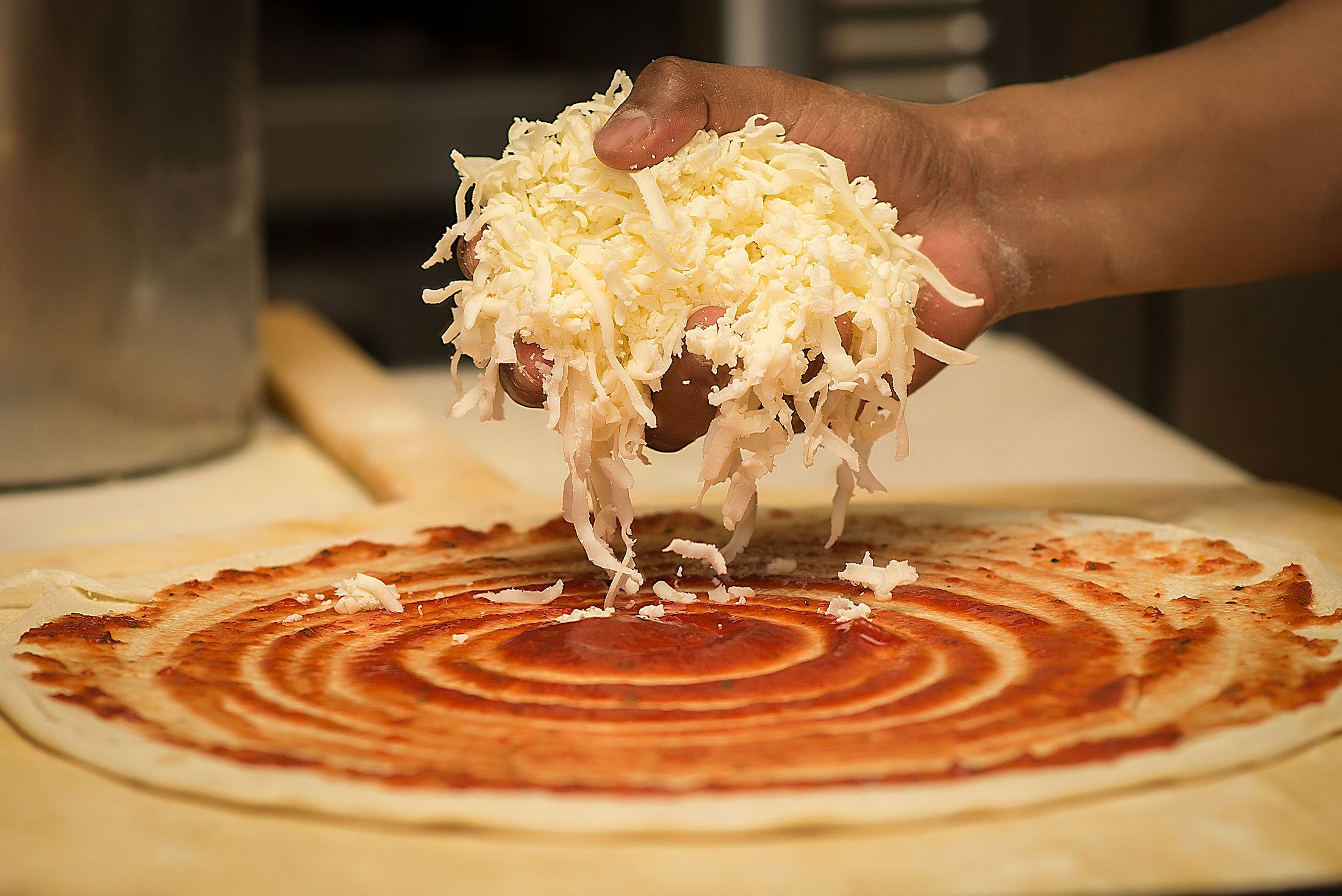 Hand sprinkling shredded cheese over pizza dough with tomato sauce.