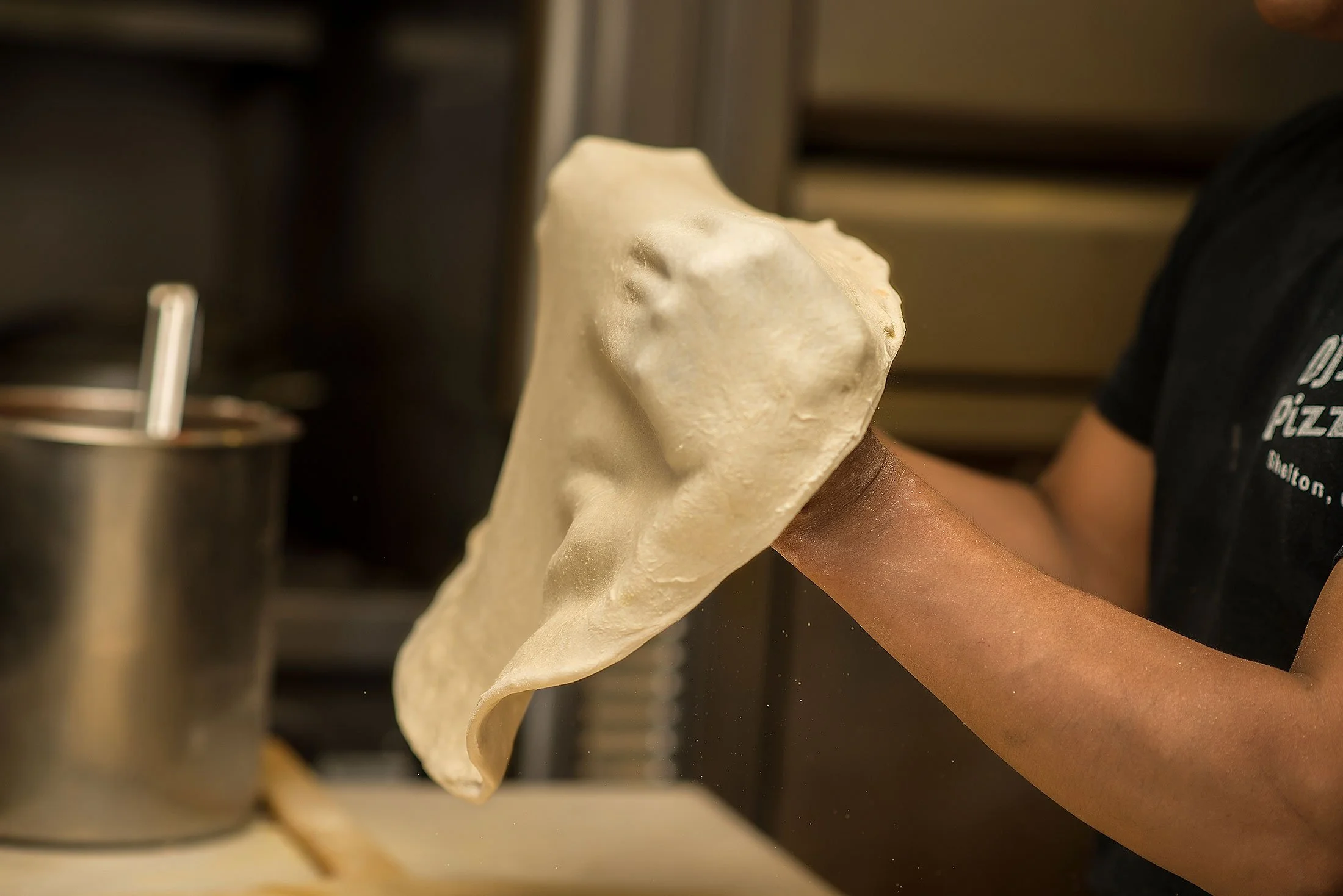 A person tossing pizza dough in a kitchen, with a metal container in the background.