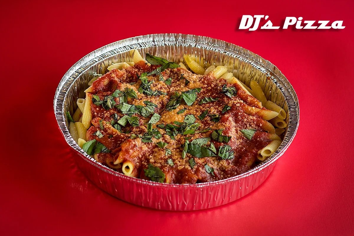 Ready-to-eat baked ziti pasta in aluminum foil container with tomato sauce, cheese, and chopped herbs on top, on a red surface, with 'DJ's Pizza' logo in the top right corner.