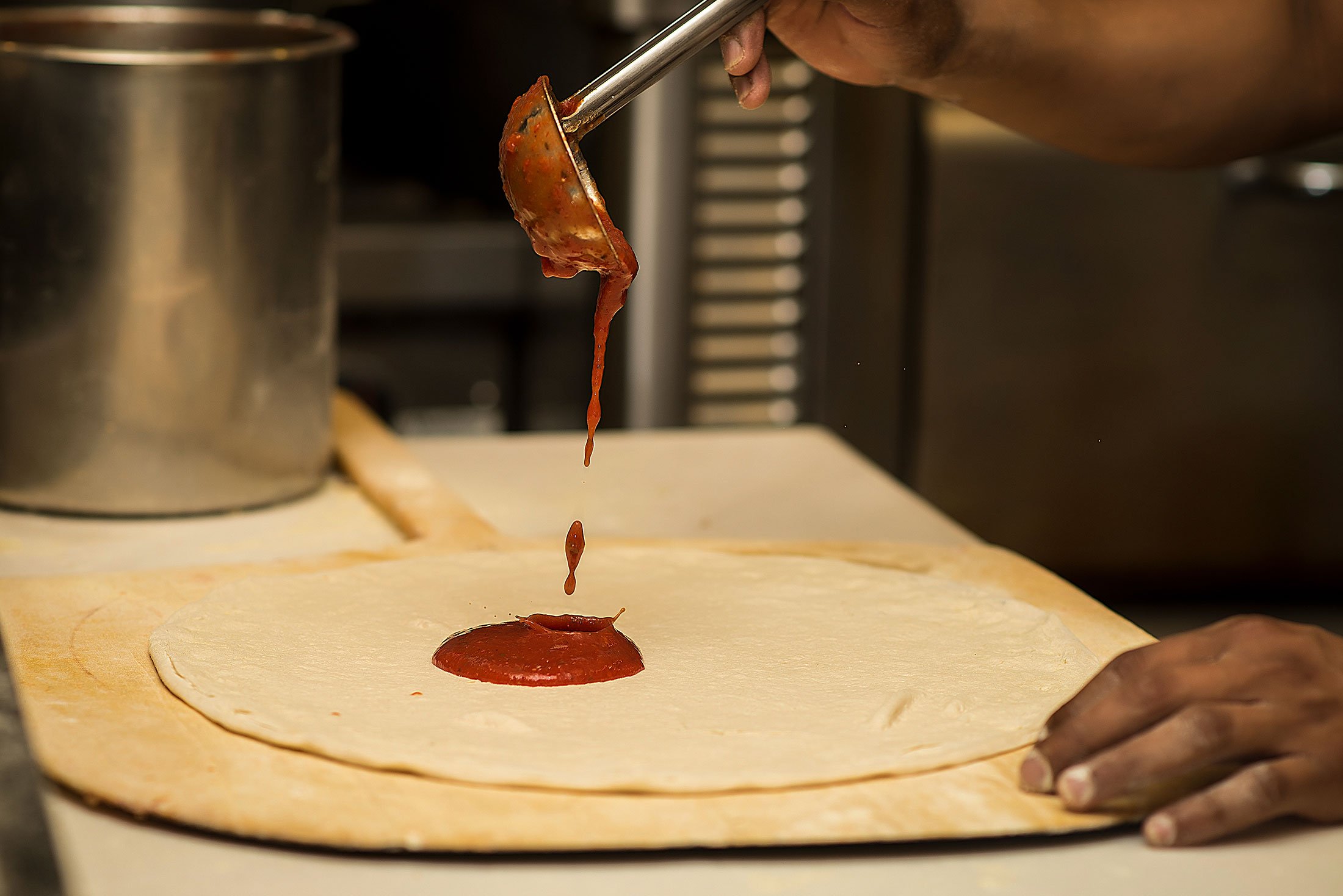 Person spreading tomato sauce onto pizza dough in a kitchen.