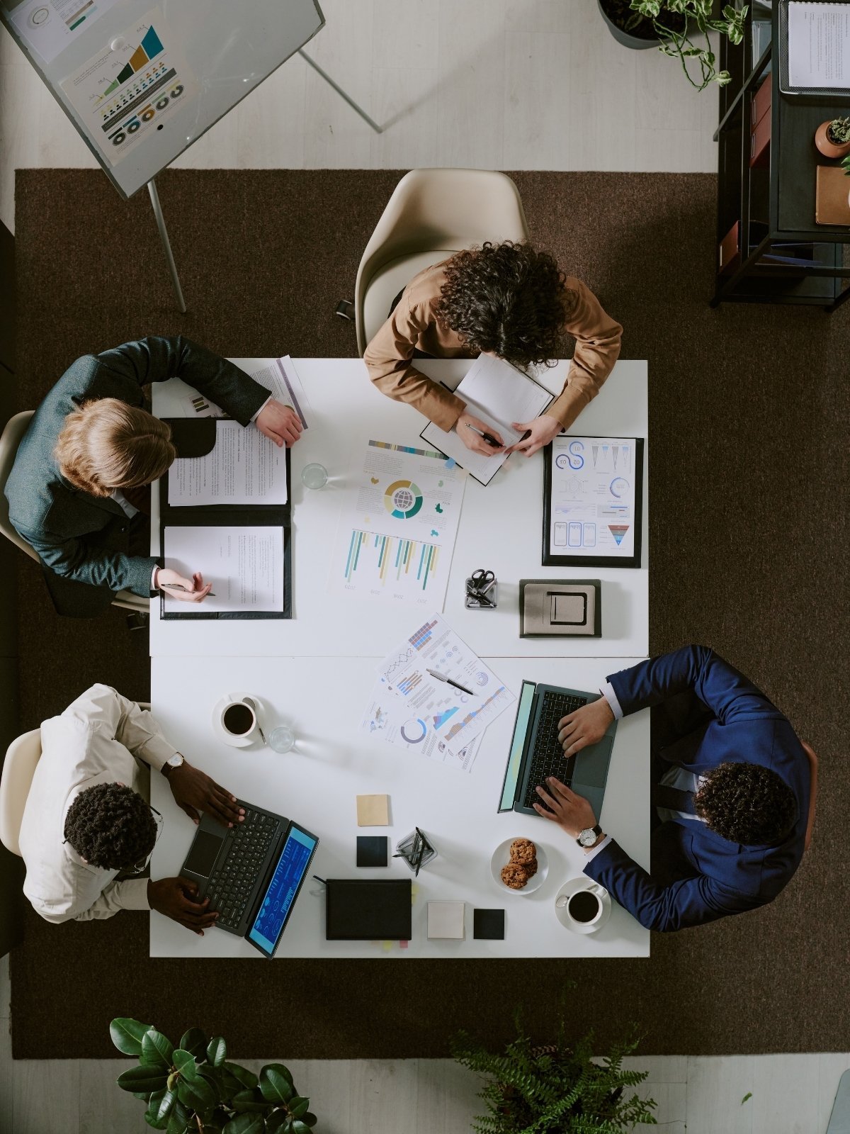 Overhead view of a business meeting with five professionals sitting around a white table. They are working on laptops, notebooks, and documents with charts and graphs. The table has coffee cups, cookies, and office supplies. The setting includes plants and office shelves.