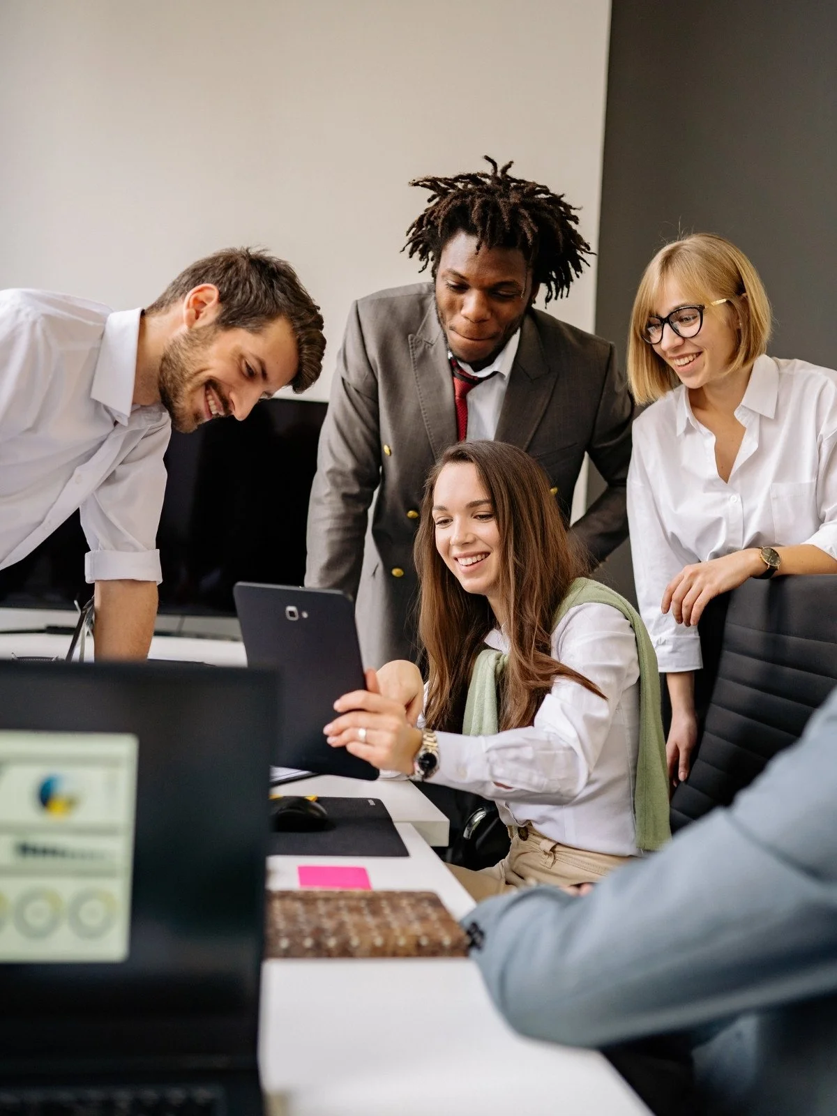 Group of five diverse young professionals looking at a tablet in a modern conference room, smiling and collaborating.