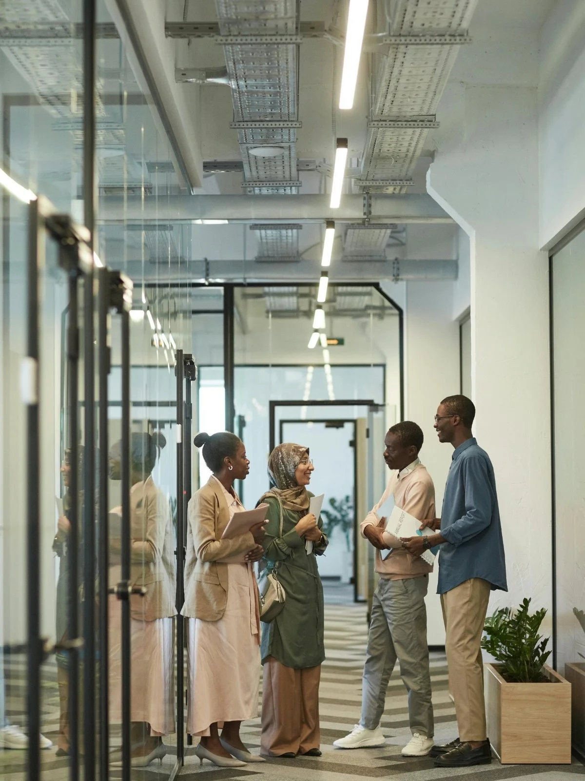 Four people talking and smiling in a modern office hallway, holding notebooks and papers.